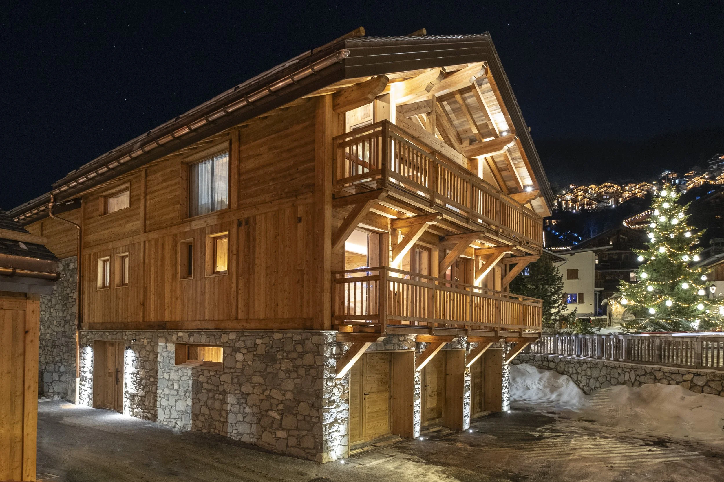 Nighttime view of a three-story wooden chalet with stone foundation, lit from inside, with balconies and a Christmas tree decorated with lights nearby.