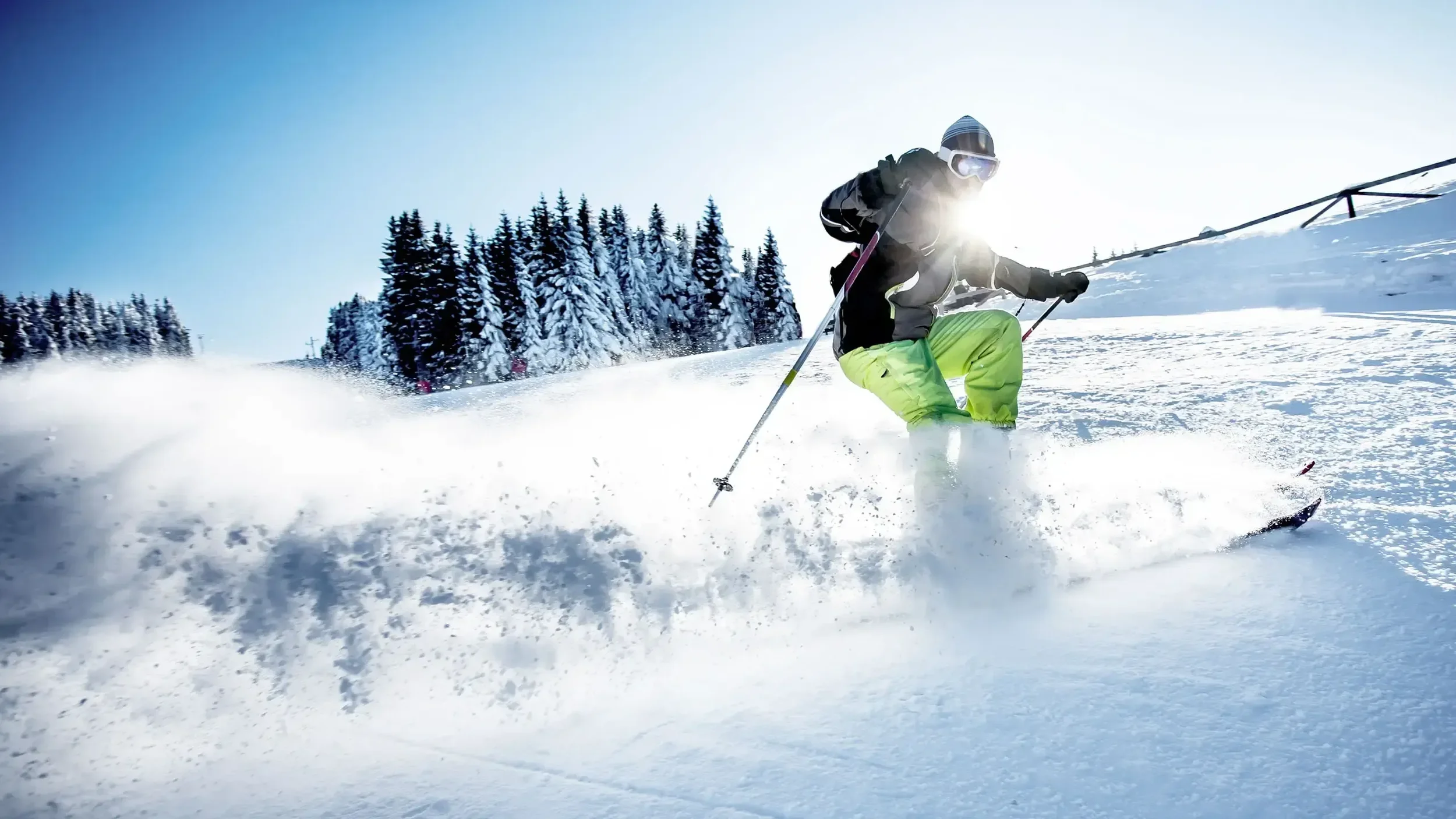 Skier with green trousers, black jacket, hat and goggles doing a hockey stop spraying snow.