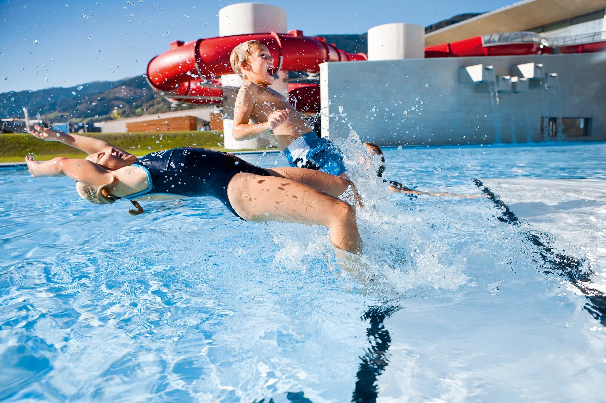 Children playing in a swimming pool near a water slide, with a water park building and mountains in the background.