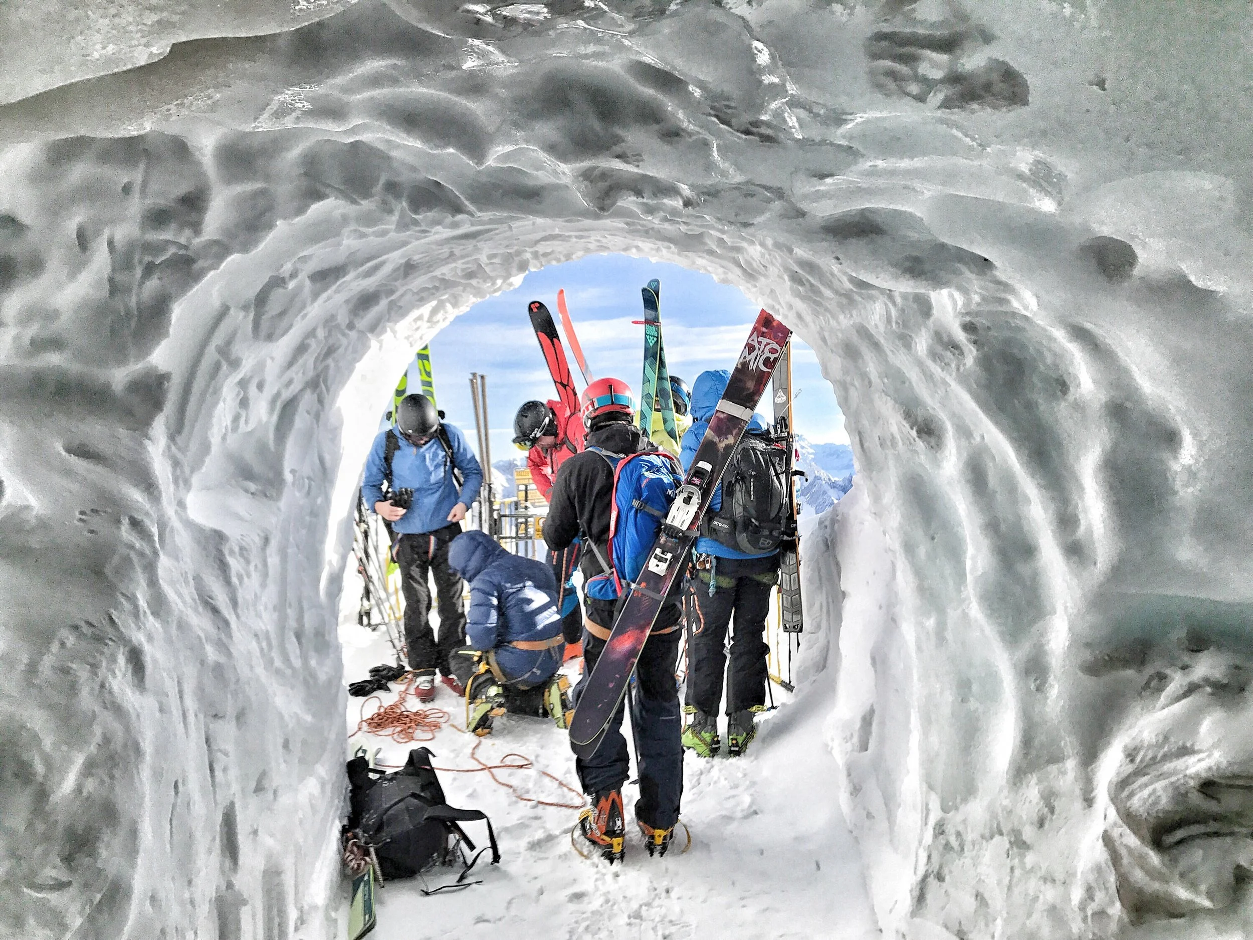 Skiers and snowboarders preparing at the entrance of an ice tunnel on a snowy mountain.