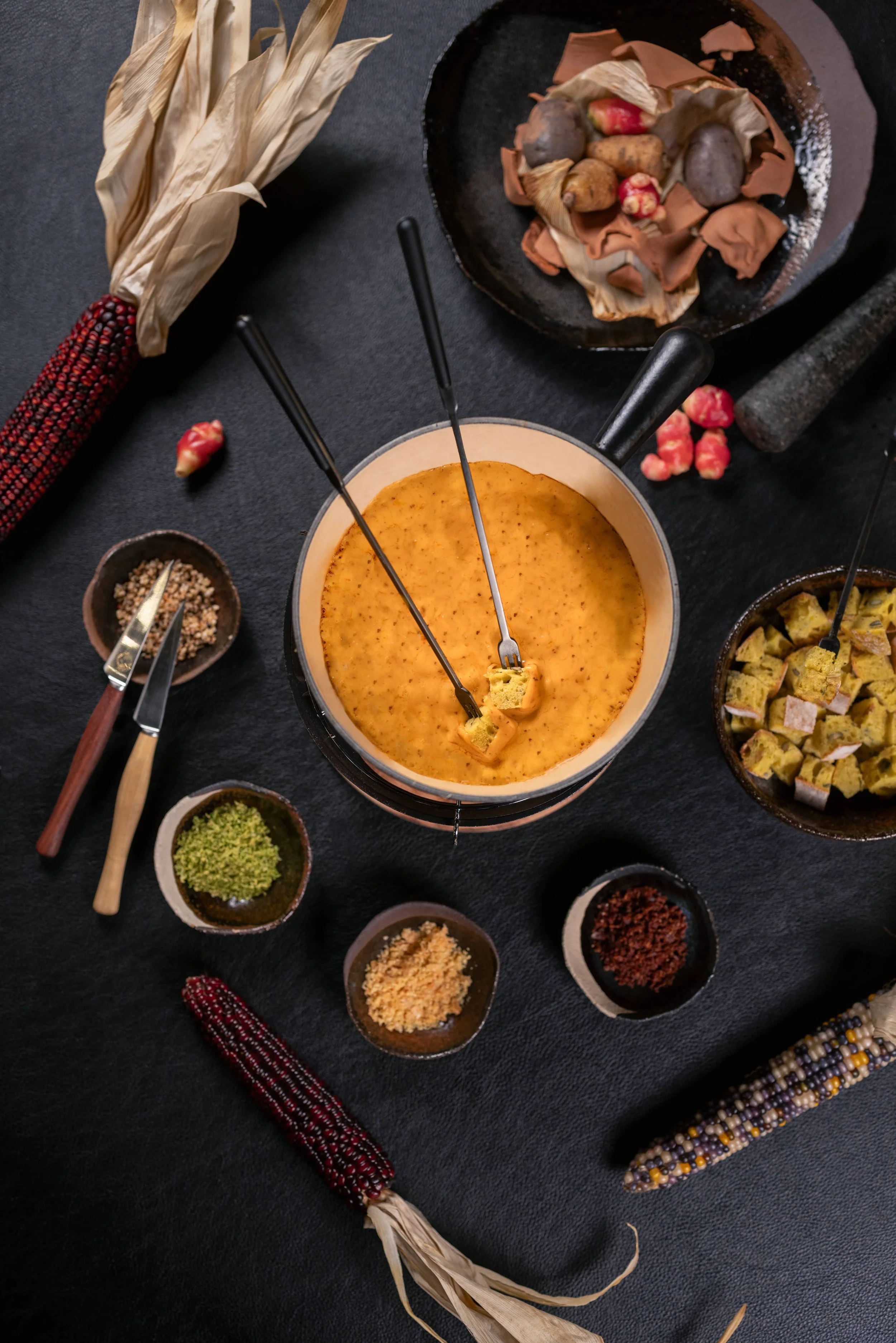 A cheese fondue in a pot surrounded by small bowls of spices, seasonings, and dippers such as bread cubes, vegetables, and berries, with decorative dried corn cobs around the setup.