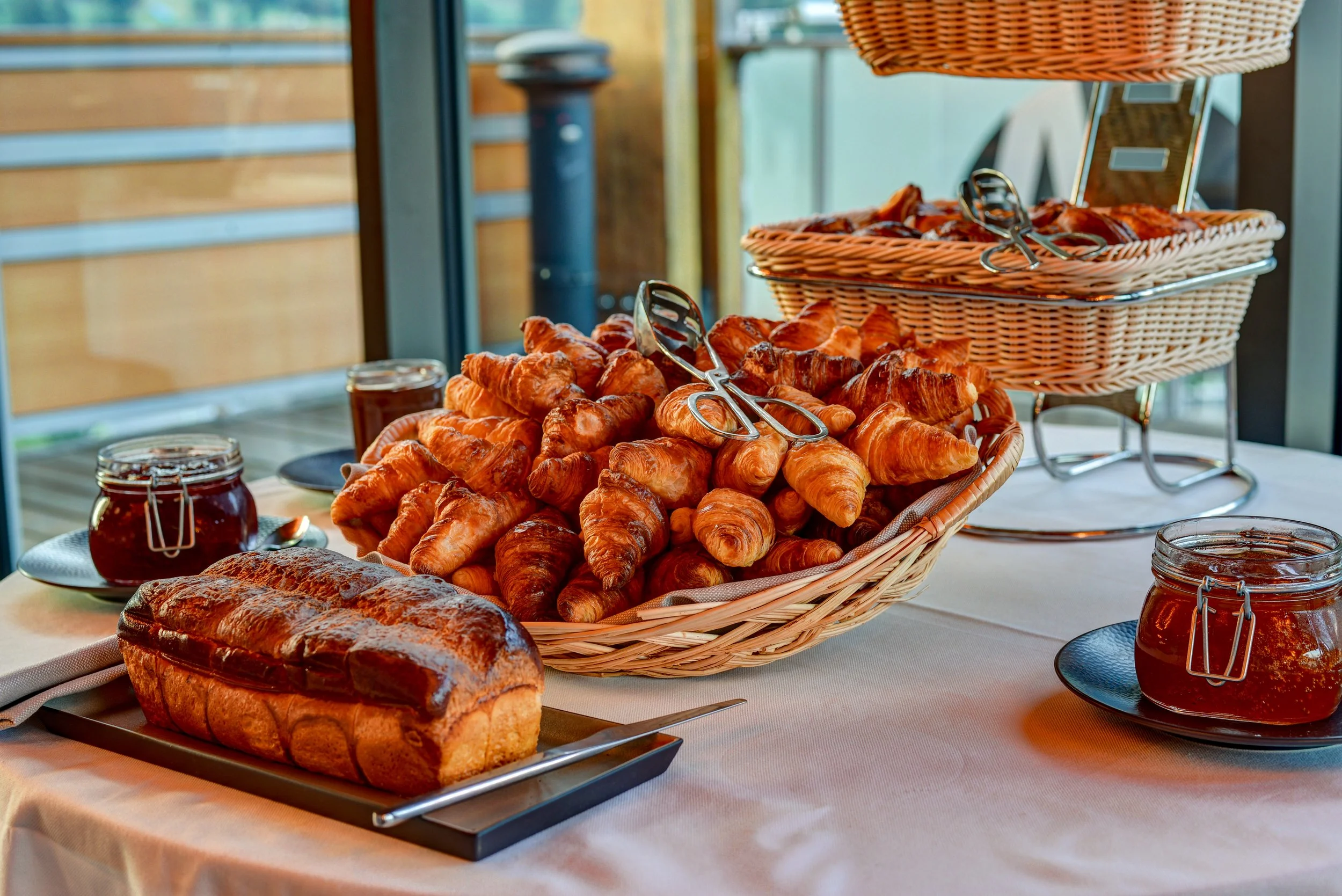 A breakfast buffet with croissants, loaf bread, and jars of jam on a table.