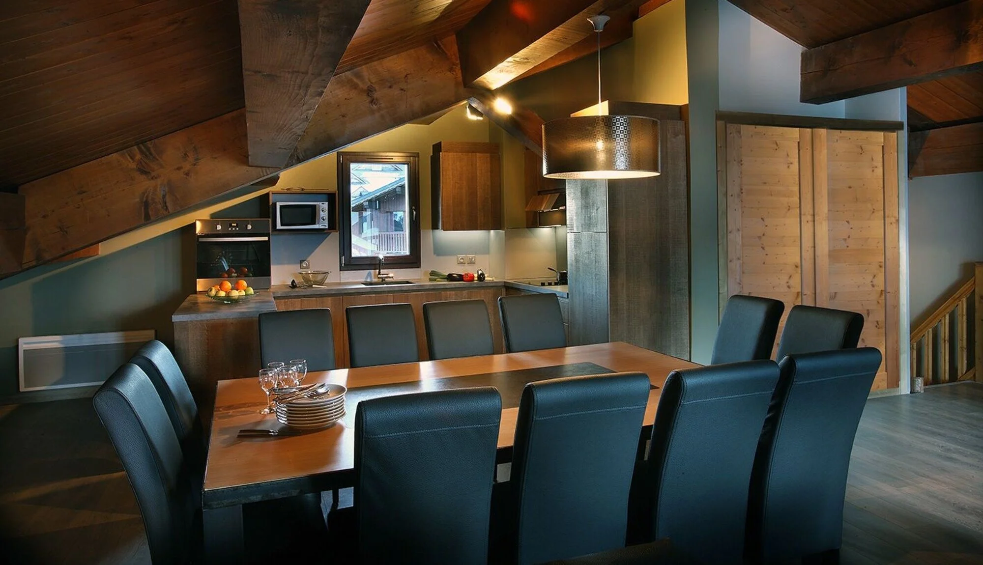 Black leather chairs around a wooden table. Open plan kitchen behind. Subtle lighting and atmospheric.
