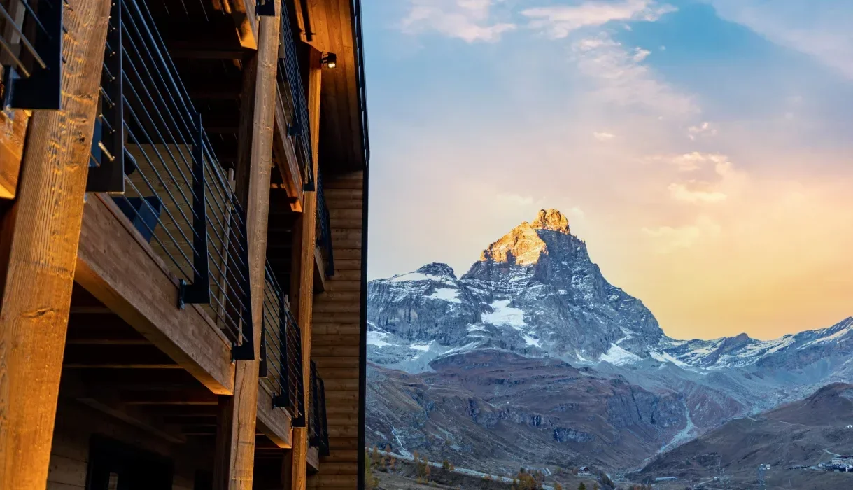 A mountain with snow on top and rocky slopes, visible during sunset or sunrise, with part of a wooden building with balconies in the foreground.