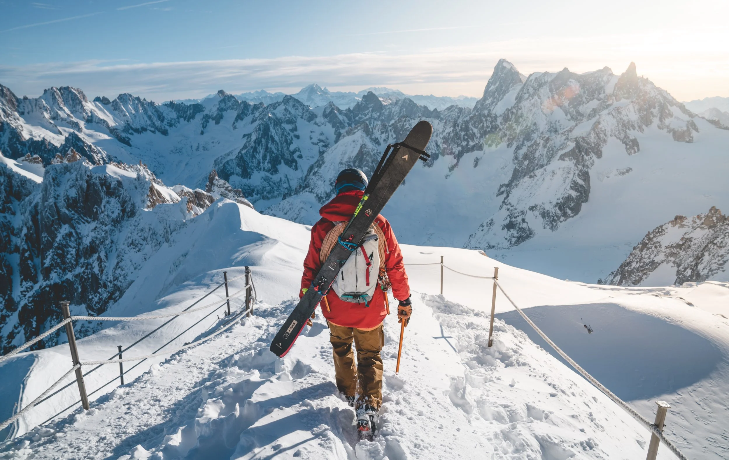 A person in winter gear walking along a snow-covered mountain trail with skis strapped to their back, overlooking a snowy mountain range under a clear sky.