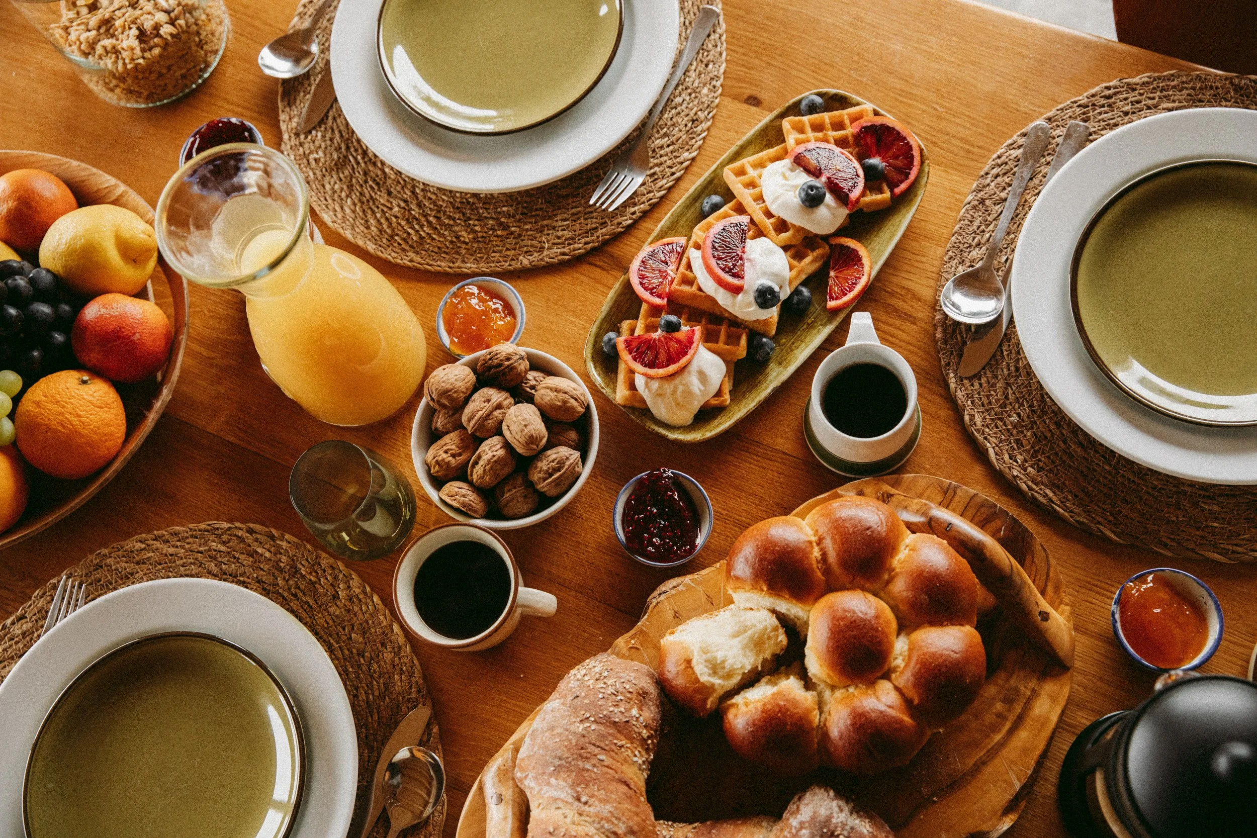 A breakfast table with plates of green soup, bread rolls, a plate of fruit, waffles with fruit and whipped cream, nuts, jam, and breakfast beverages including orange juice and coffee.