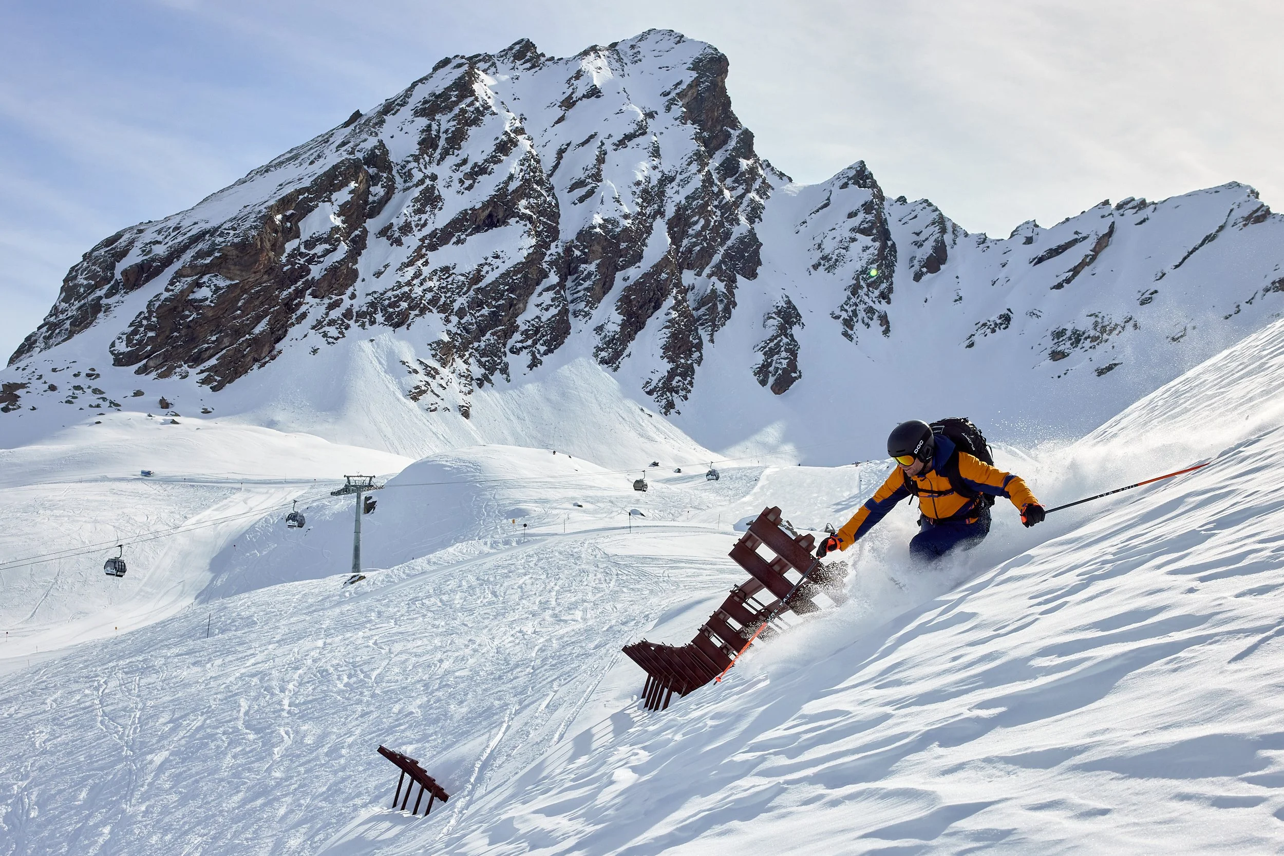 Orange jacket skier skis down the side of the mountain in fresh powder conditions on a sunny day.