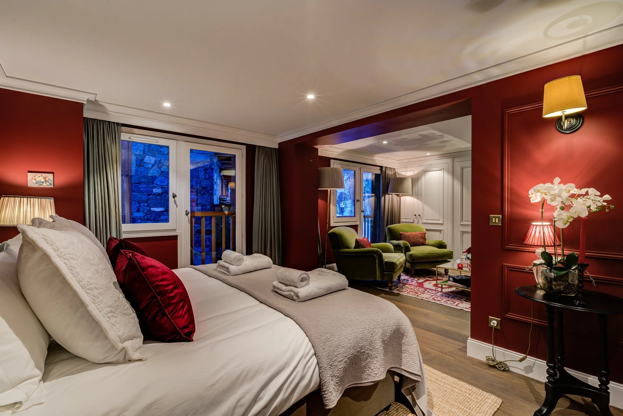 Double bedroom suite with red walled interior, British style with light vaulted ceilings. A remarkable bedroom with a living area of green arm chairs.