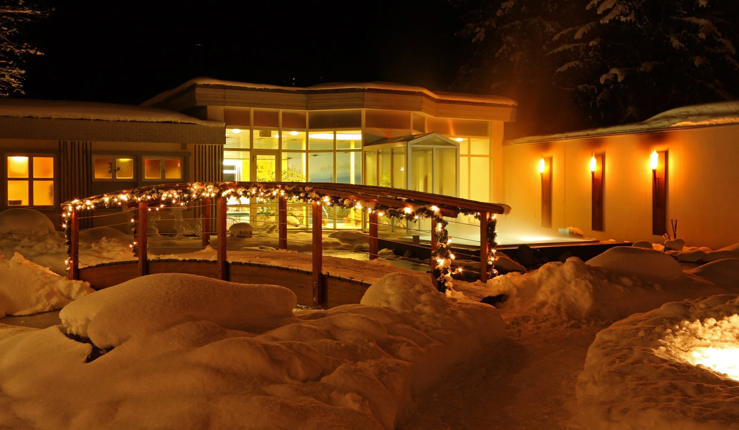 A snowy outdoor scene at night with a building illuminated by warm yellow light. A snow-covered wooden bridge decorated with holiday lights crosses a snow-covered path. The building has large glass windows and wall-mounted lights.