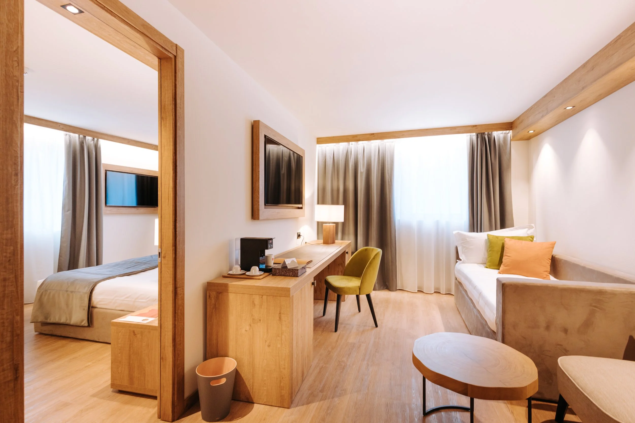 A modern hotel room with a bedroom and a sitting area. The bedroom has a bed with beige bedding and a window with curtains. The sitting area features a sofa with colorful pillows, a small table, and a TV mounted on the wall. There is wooden furniture including a desk, a chair, and trim around the window and doorways.