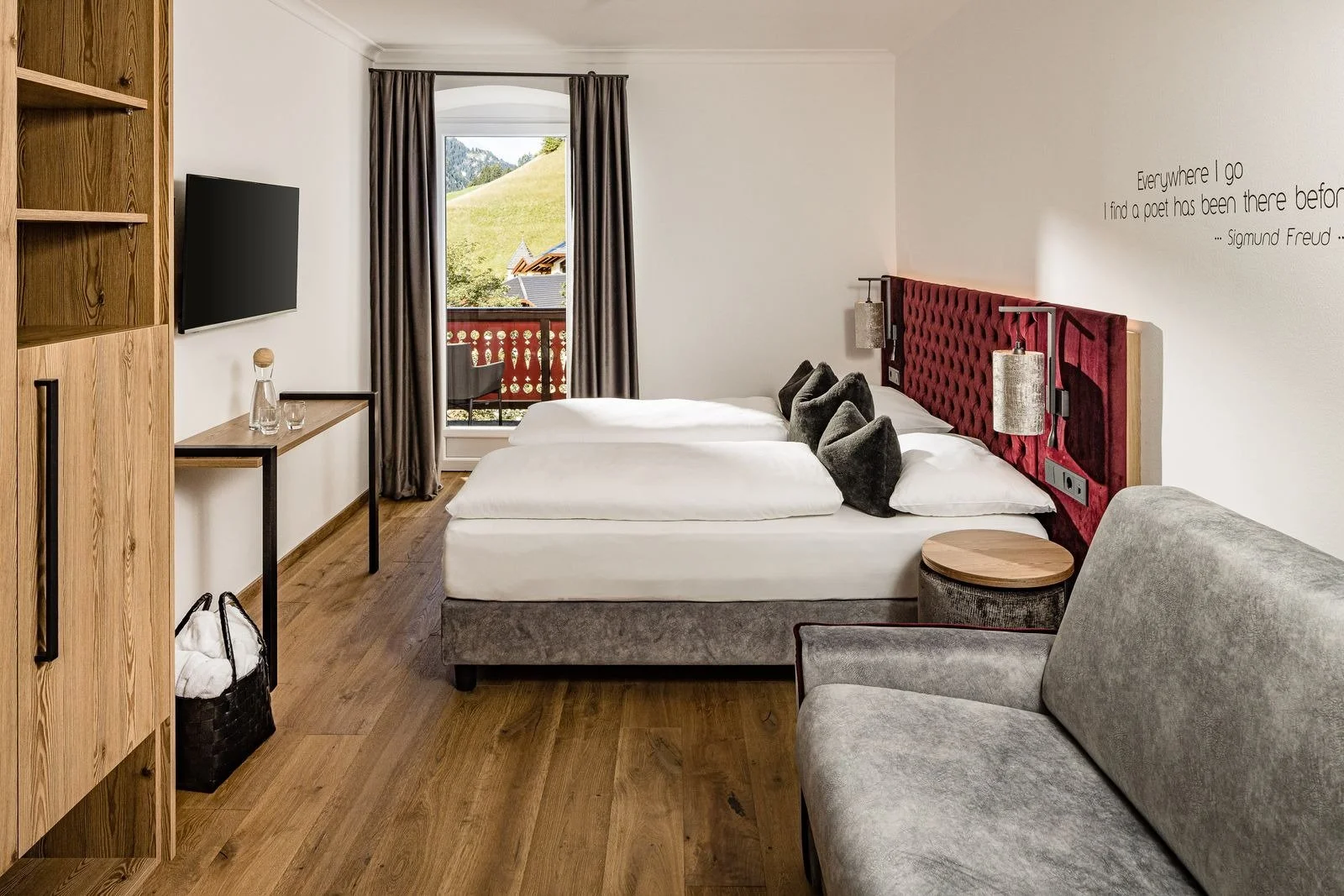 Double bedroom with grey sofa, red headboard and white bed. Sights of the Italian mountains in the background.