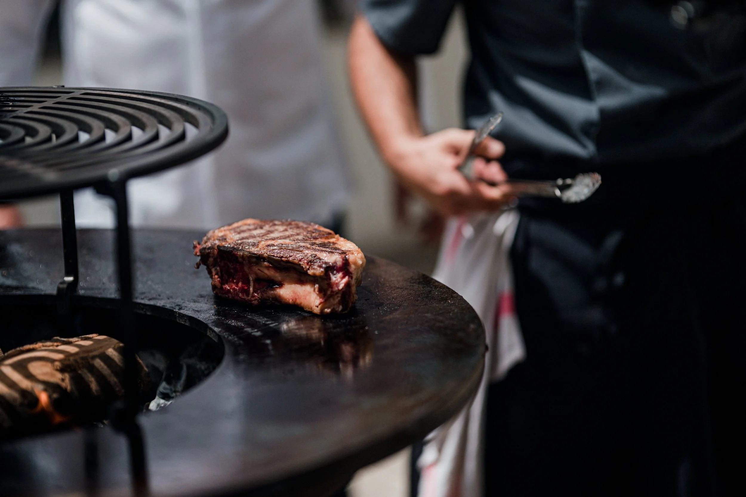 A piece of grilled steak on a black grill surface with a person in the background holding tongs, wearing a black shirt, in a restaurant or kitchen setting.