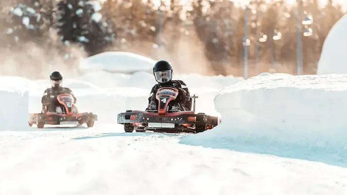 Two young children racing in go-karts on a snowy track with snow banks on sides, winter trees in the background, and sunlight coming through.