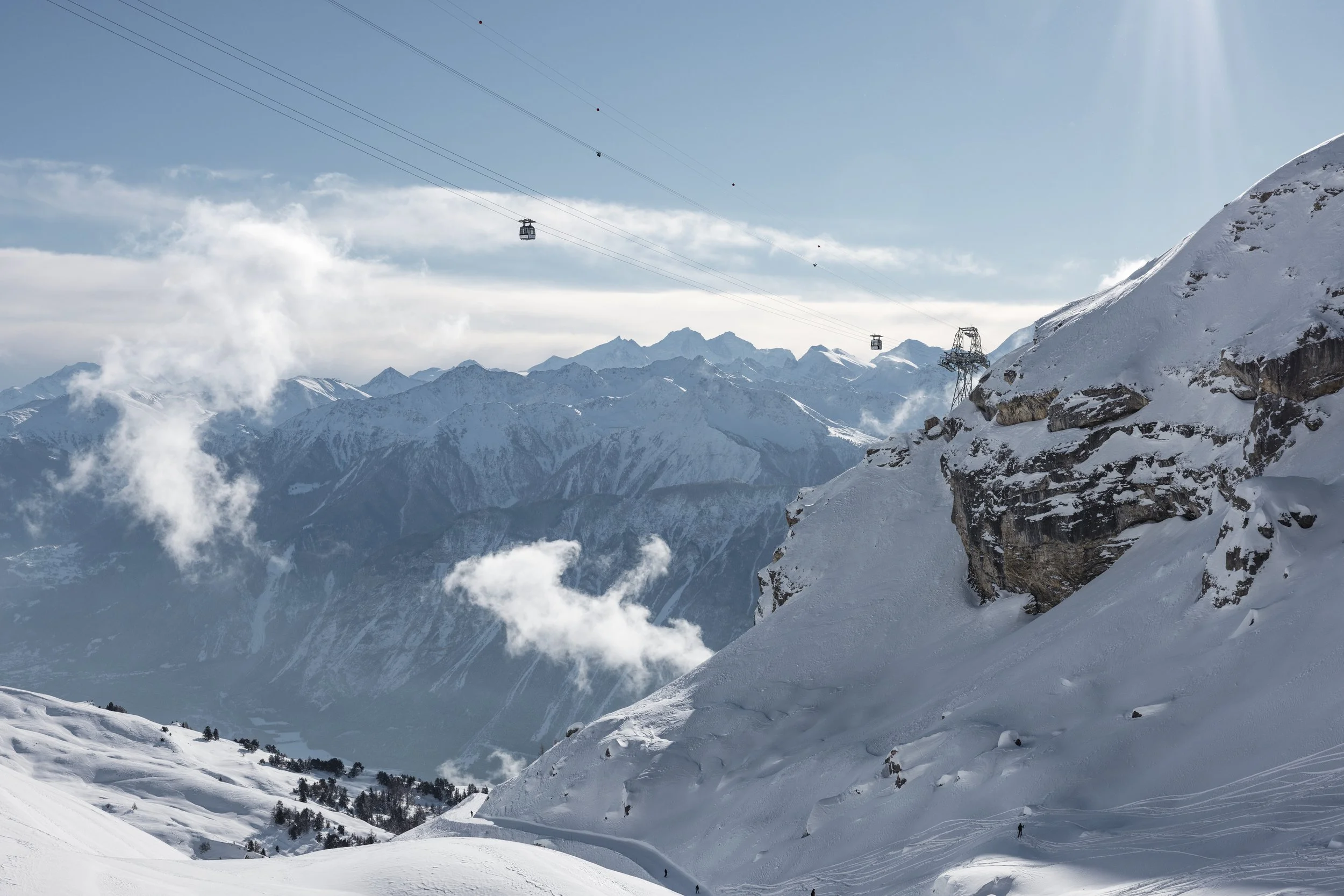 Snow-covered mountain landscape with ski slopes, mountain range in the distance, ski lift cables overhead, and blue sky with some clouds.