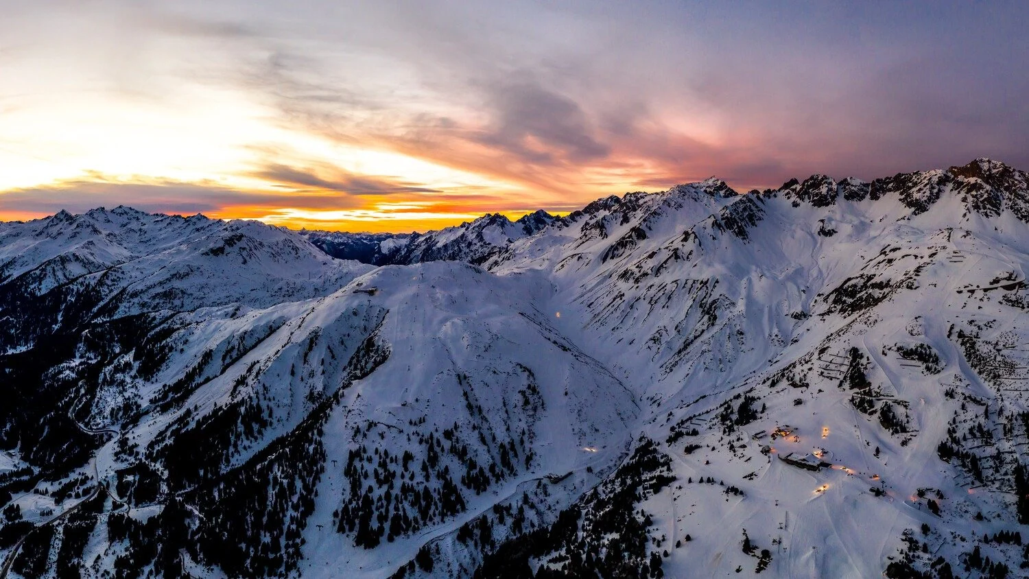 Snow-covered mountain range at sunset with a colorful sky and small illuminated buildings scattered across the slopes.