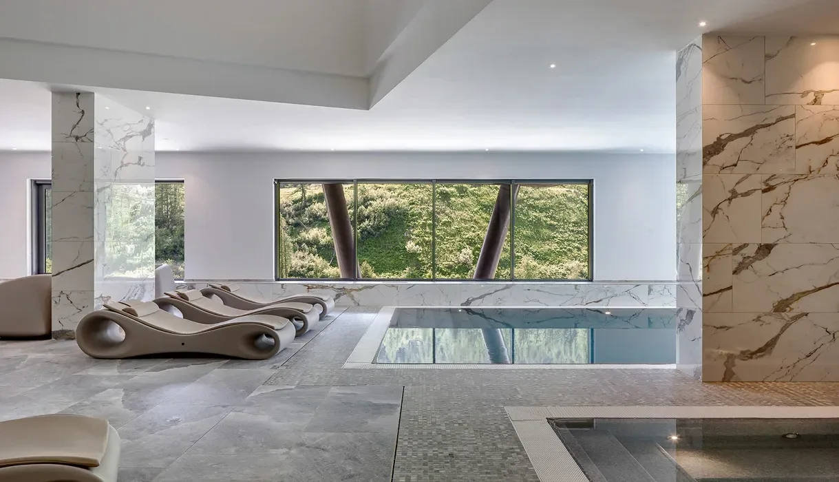 Indoor pool area with modern lounge chairs, marble walls, and large windows overlooking greenery.