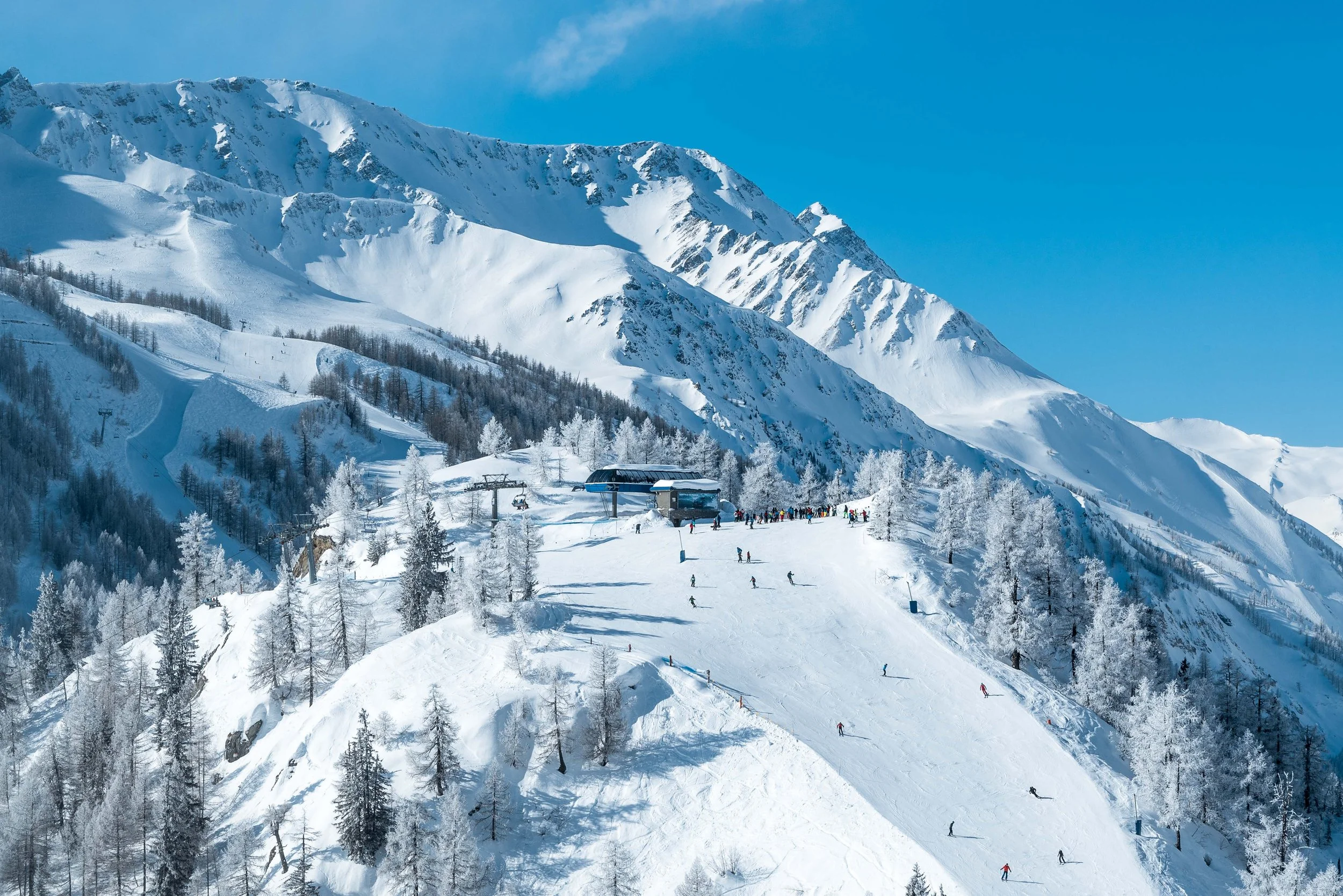 Ski resort on snowy mountain with skiers, snow-covered trees, lift, and blue sky.