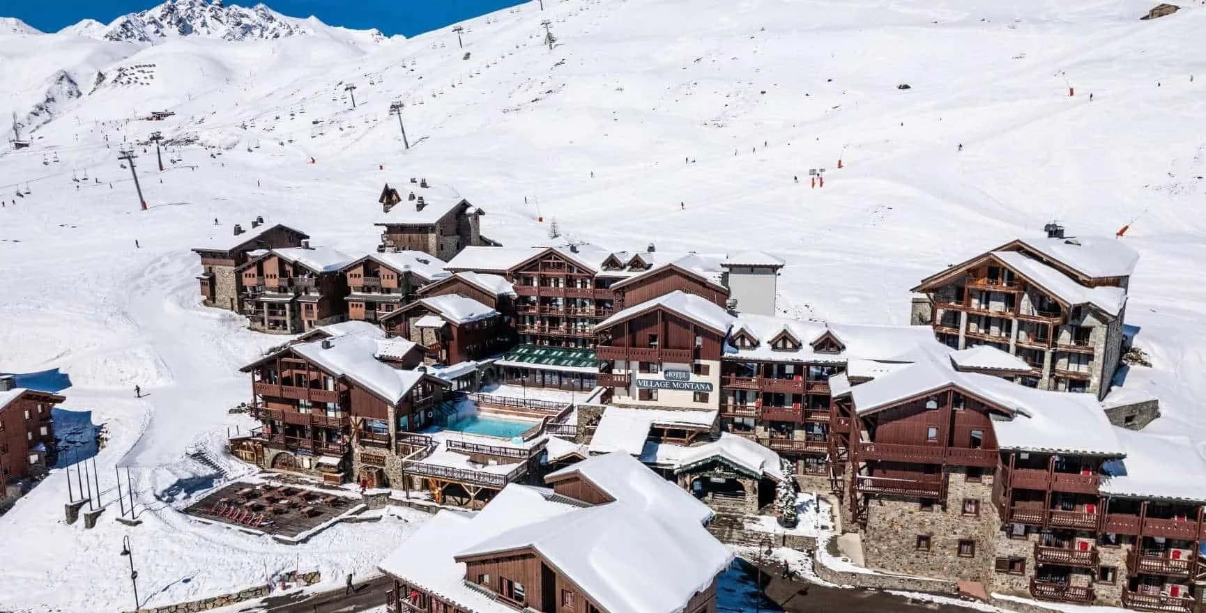 Ski resort village with multiple chalet-style buildings with snow on their roofs, a swimming pool, ski slopes in the background, and ski lifts on the mountainside.