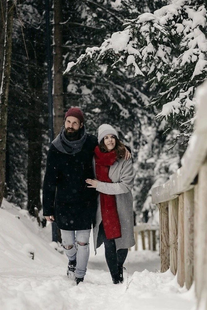 A man and woman walking together in a snowy forest during winter, with snow-covered trees and a wooden fence on the side.