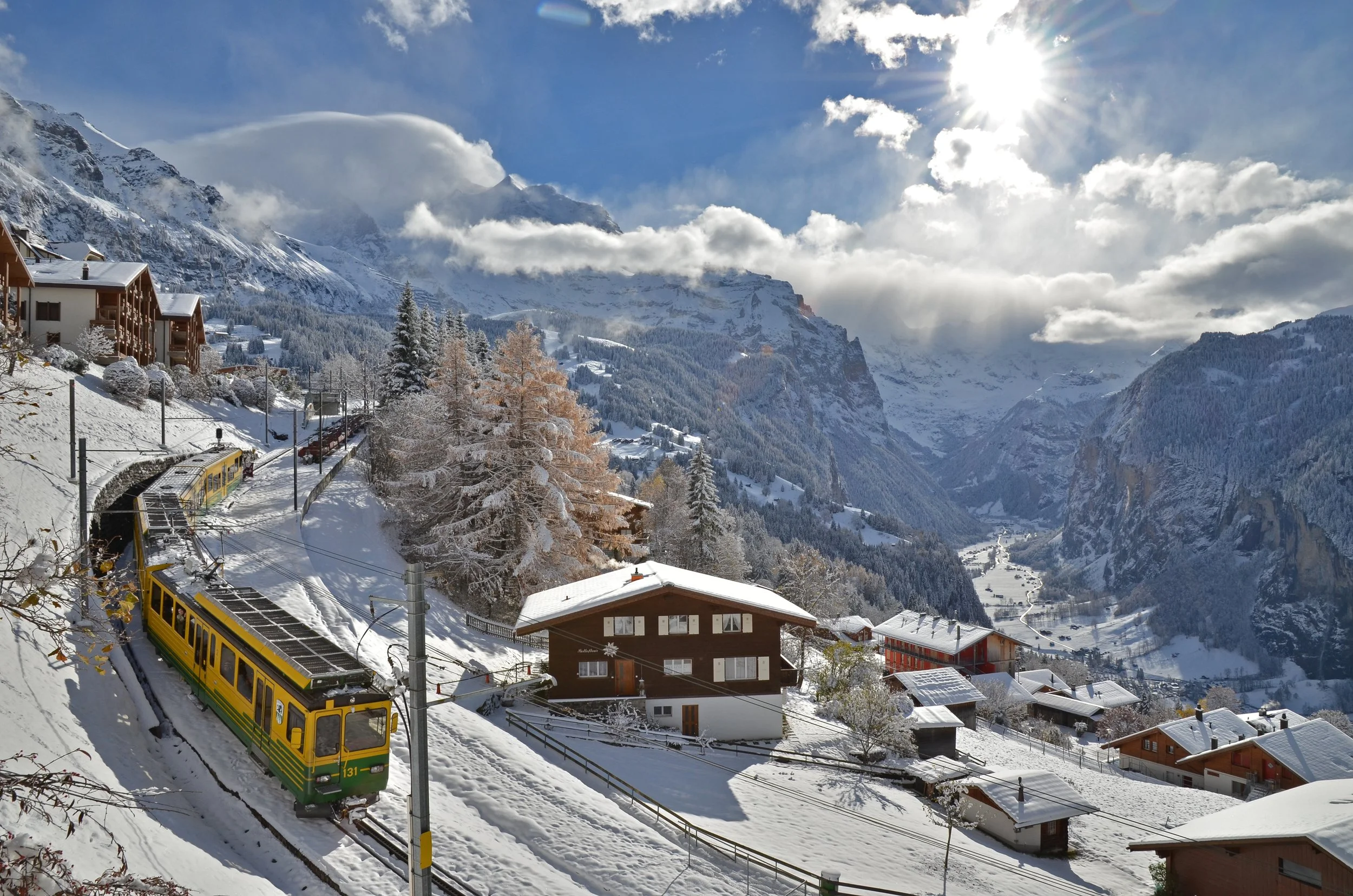 Snow-covered village in a mountainous landscape with a yellow train on a curved track, wooden houses, tall snow-laden trees, and a bright sun in a partly cloudy sky.