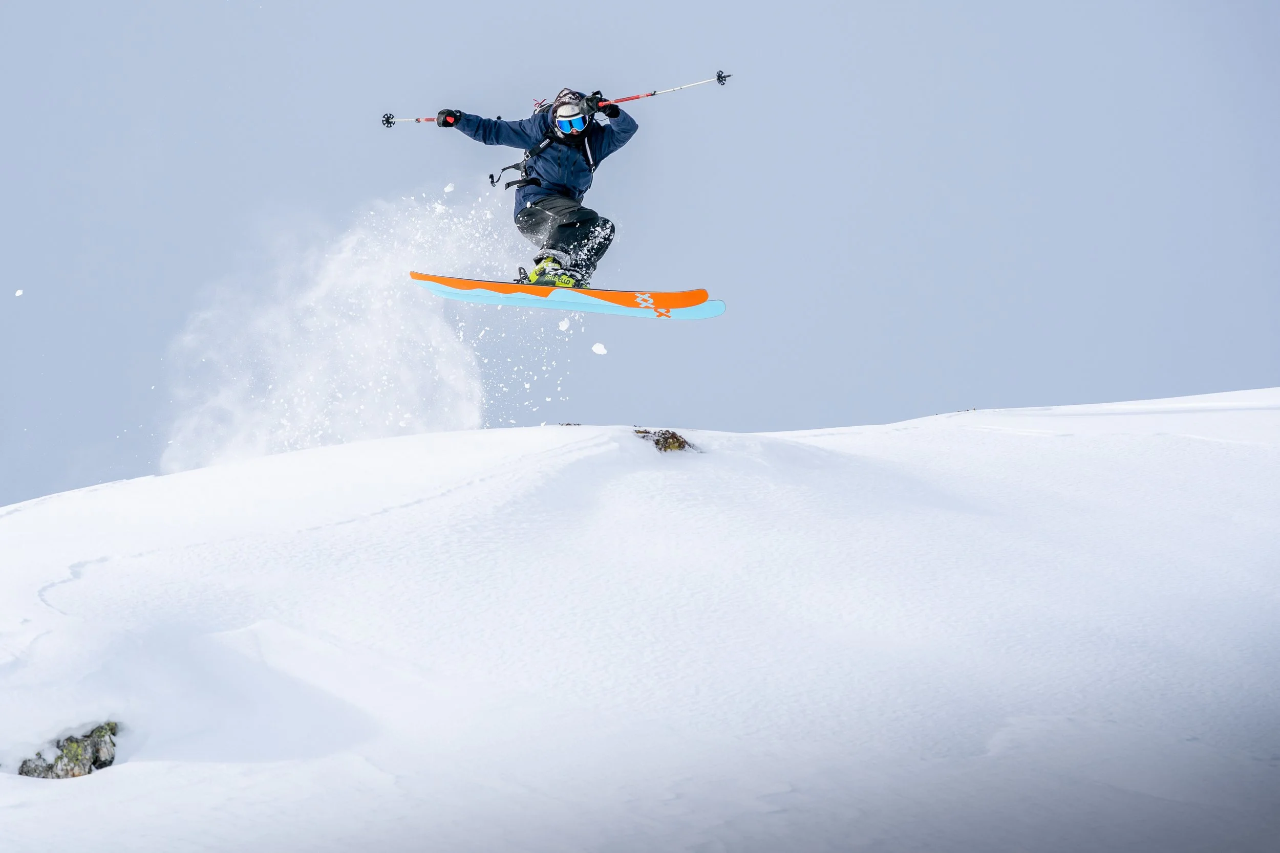 A person wearing a blue jacket, black pants, a helmet, and goggles snowboarding in mid-air above snow-covered terrain with a cloudy sky.