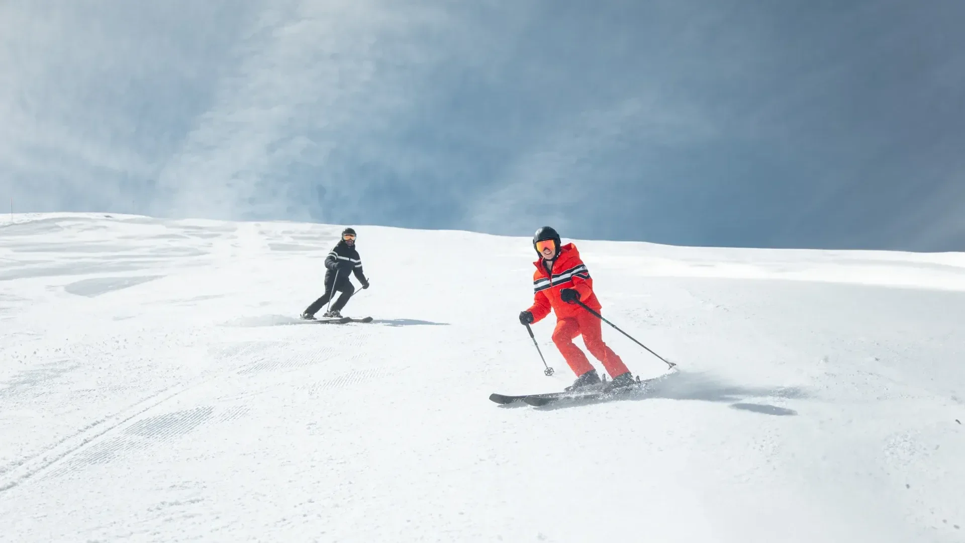 Two intermediate skiers make their way down the mountain. One looks to be the instructor with blue skies above.