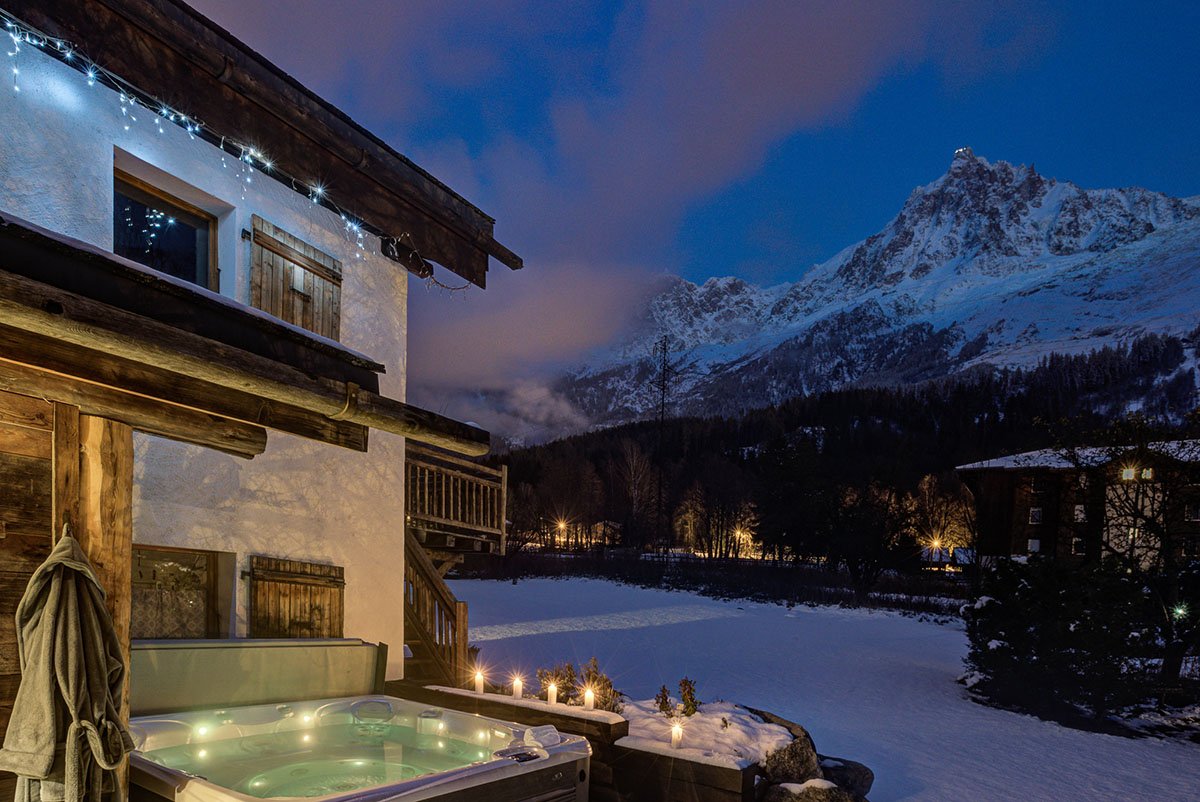 A hot tub on a wooden deck outside a chalet with snowy mountains in the background during winter evening.