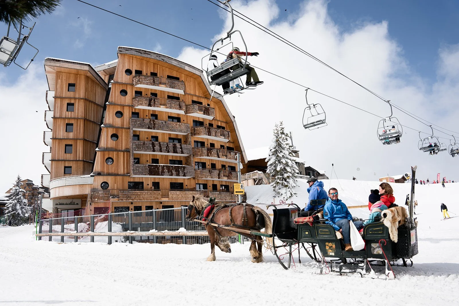 People riding in a horse-drawn carriage on snow in front of a modern wooden building at a ski resort, with ski lifts and skiers visible in the background.