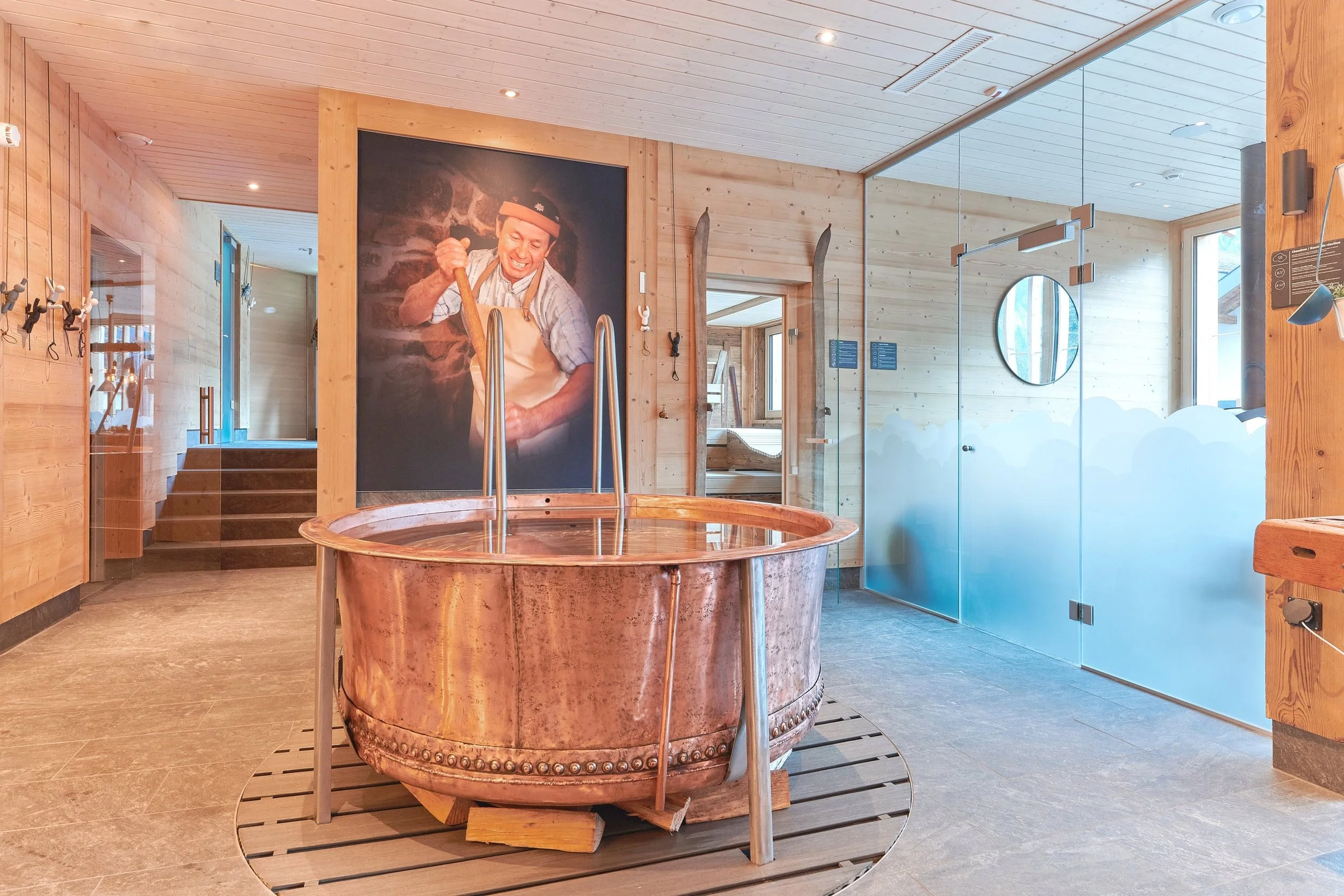 Indoor spa area with a copper hot tub, wooden walls, a large photo of a smiling man in traditional attire on the wall, and a glass door with foggy design.