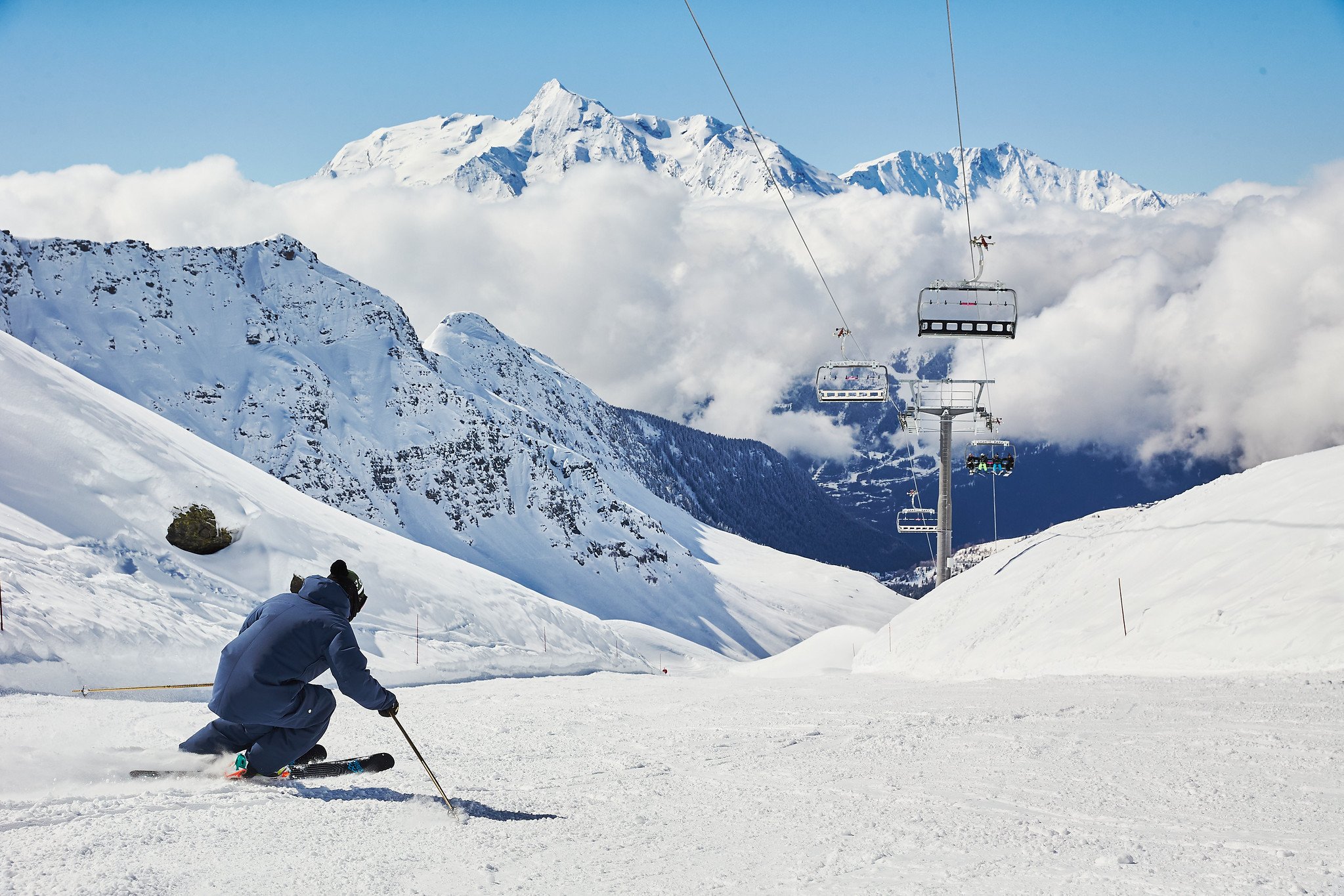 Skier heading down the mountain with a blue jacket and black skis. Ski lift overhead, blue skies and lovely views.