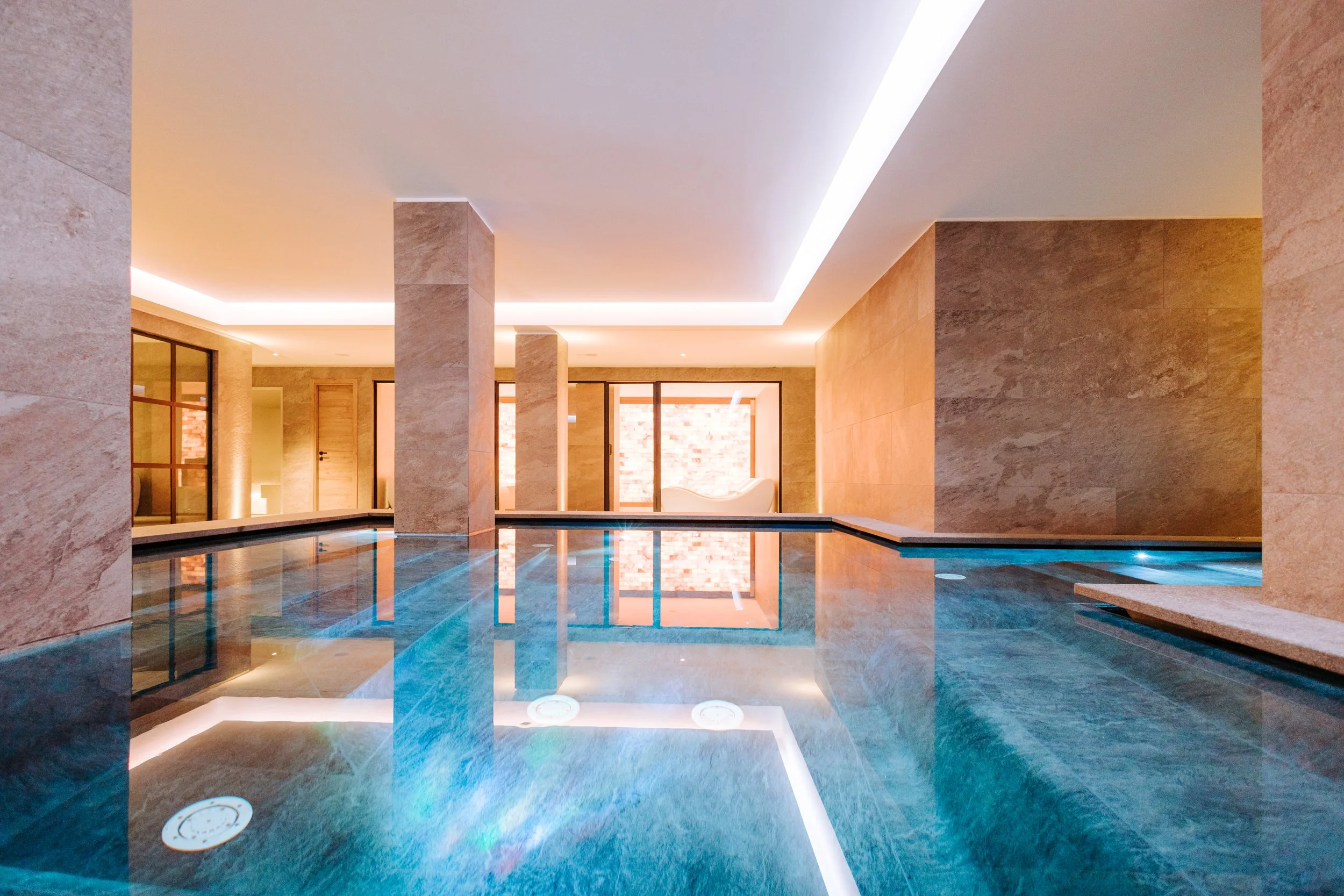 Indoor swimming pool area with beige stone walls and pillars, a white ceiling with recessed lighting, and a wide window in the background showing a brick wall.