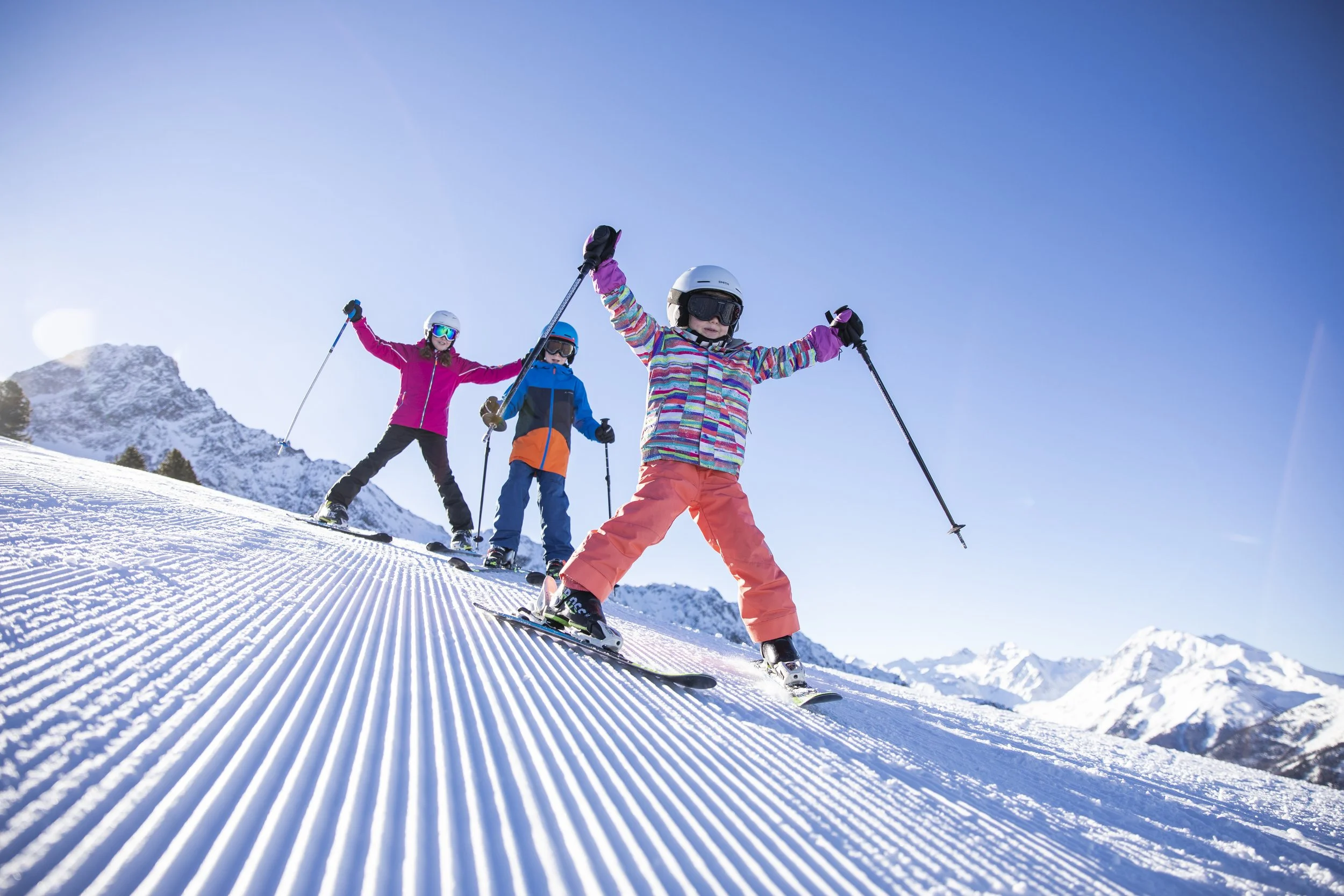 Three children skiing on a snowy mountain slope under a clear blue sky, wearing colorful ski gear and helmets.