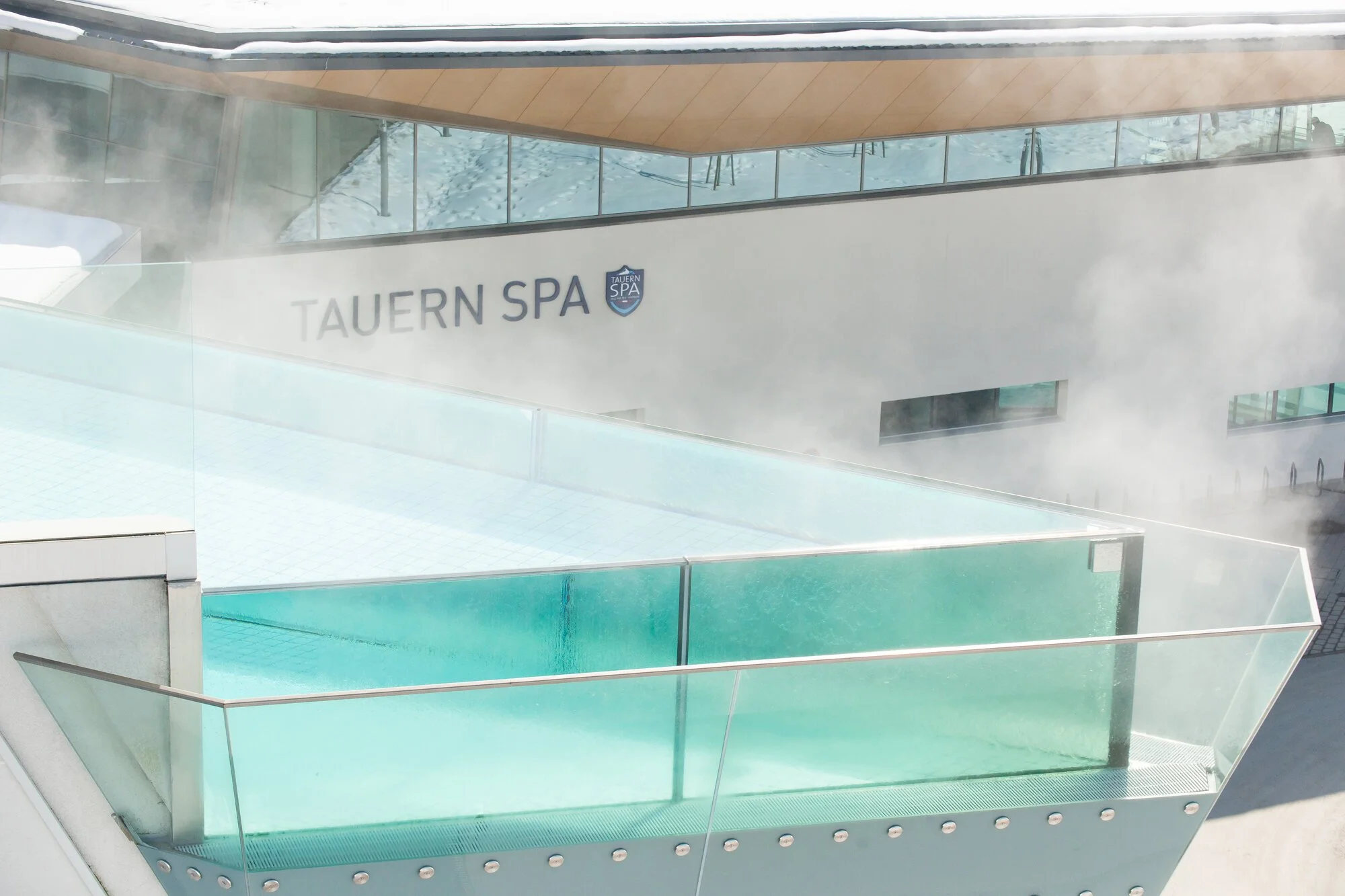 An outdoor indoor thermal hot tub or pool at Tauern Spa with steam rising from the water, glass barriers, and modern architecture.