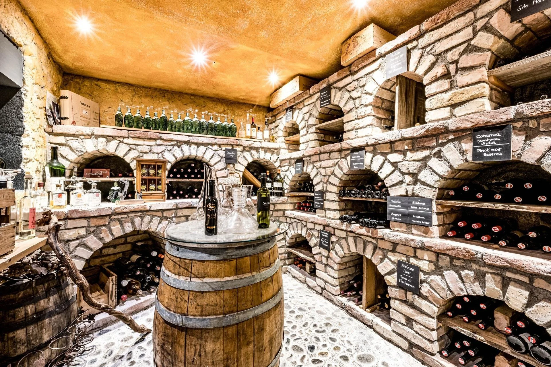 Wine cellar with brick and stone shelves filled with bottles of wine, some labels are visible. A barrel in the center holds wine bottles, glass decanters, and a wine opener, with a rustic wooden branch nearby. Small chalkboard signs label different types of wine.