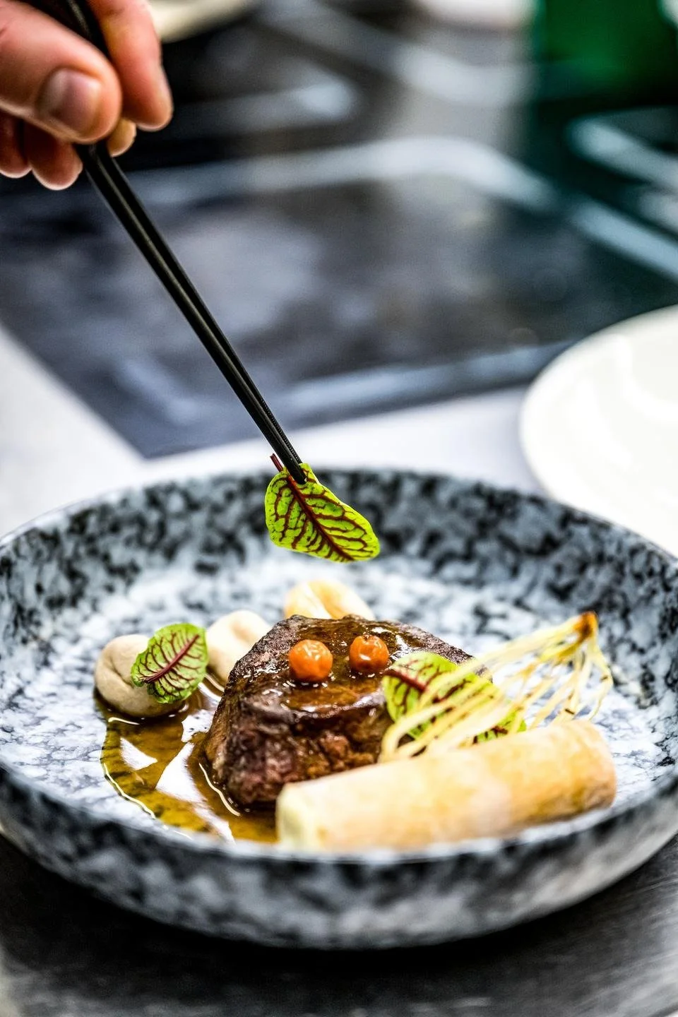 Chef garnishing a plated dish with leafy herbs using tweezers, featuring a piece of cooked meat, sauce, and vegetables on a decorative black and white plate.