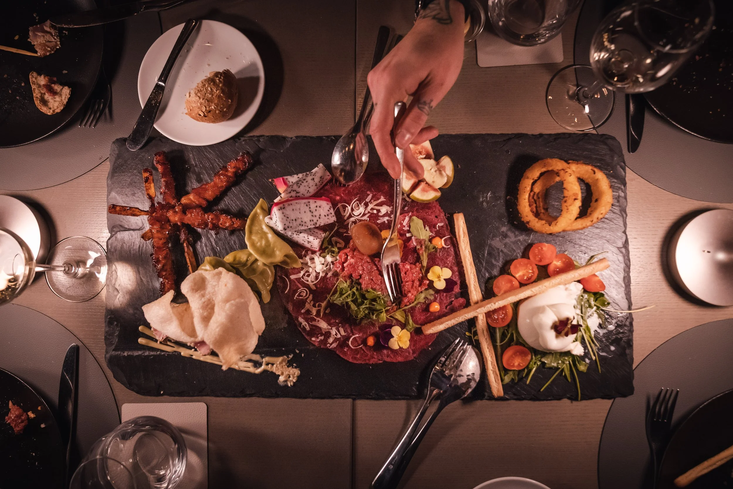 Overhead view of a slate platter with various foods, including sliced figs, cherry tomatoes, onion rings, and a dish with raw meat garnished with flowers, with a person's hand using tongs to serve. Surrounding are glasses of wine and plates with leftover food.