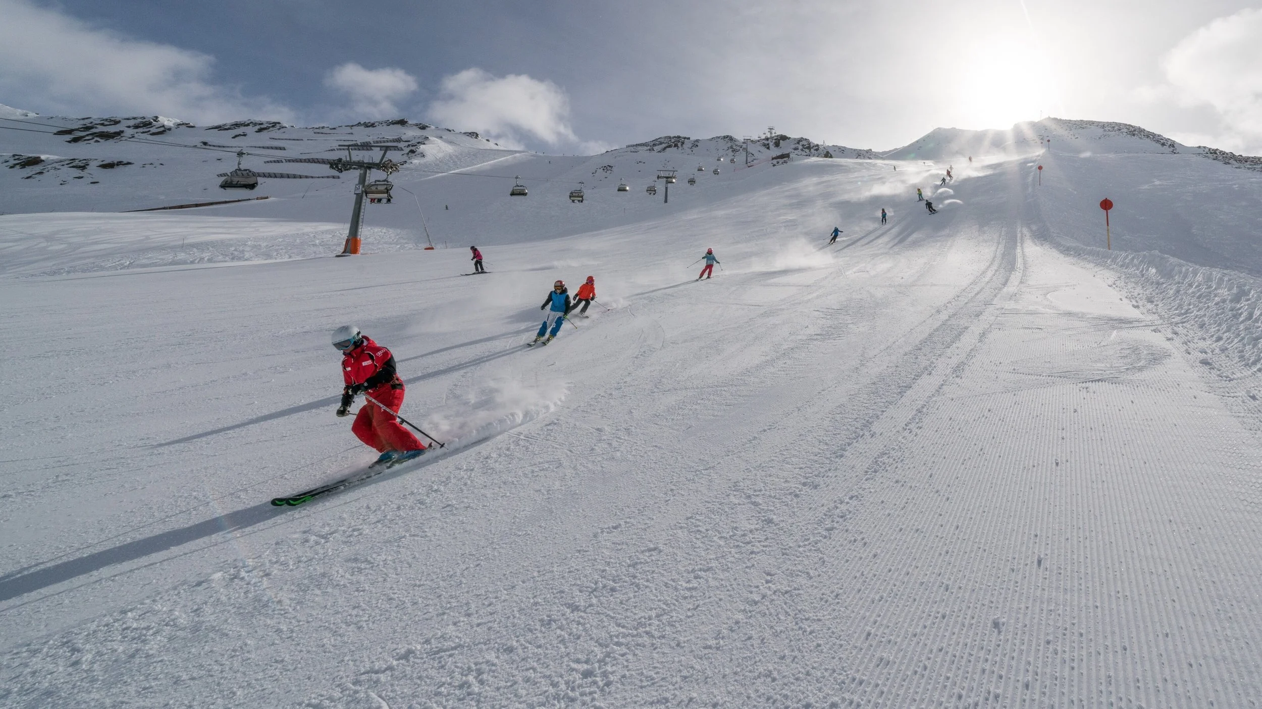 Group of people skiing down a snow-covered mountain slope on a sunny day with clouds in the sky, ski lift in the background.