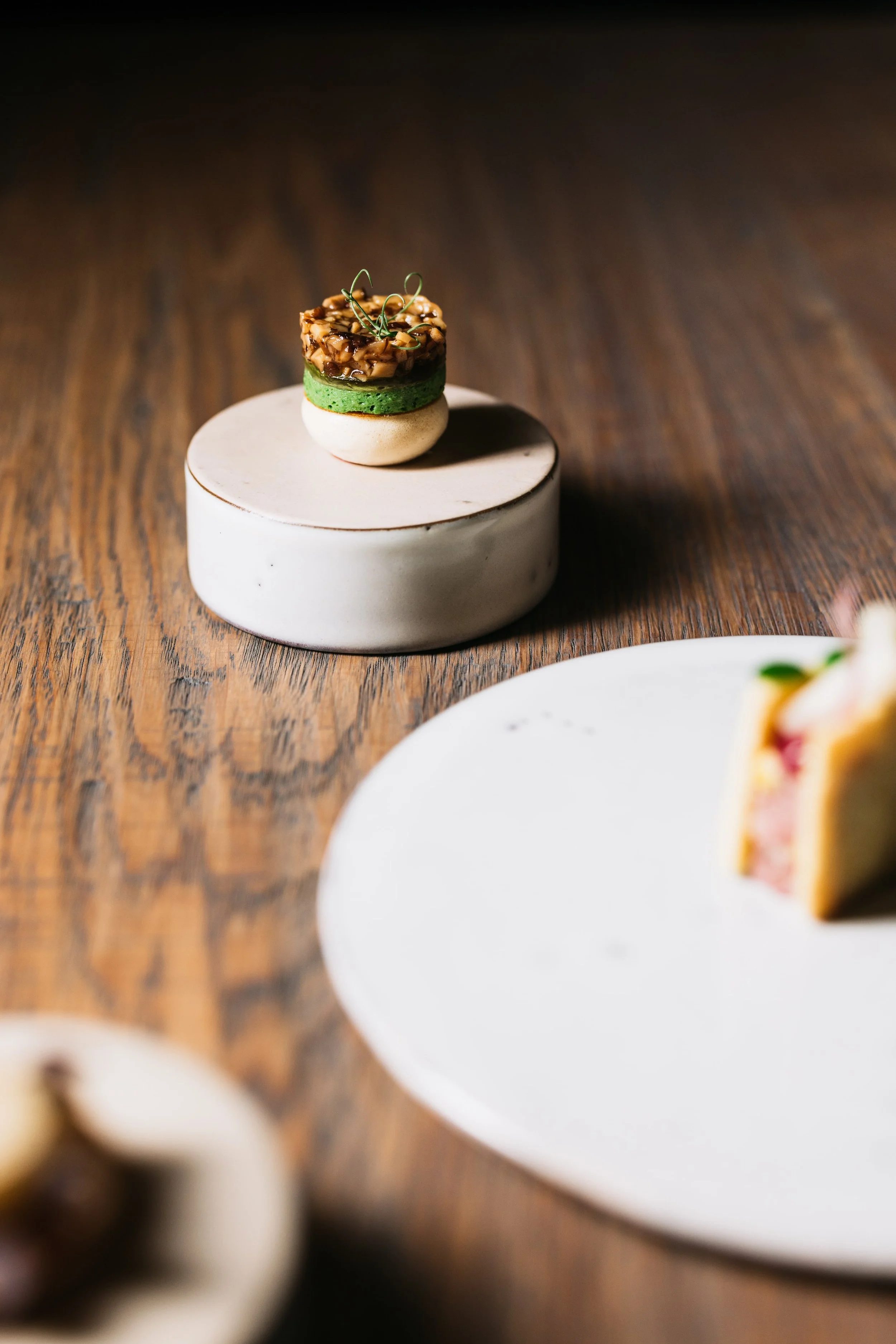 Close-up of a dessert with a layered presentation on a small white pedestal, featuring a green layer, topped with a nut and caramelized garnish, on a wooden surface with other blurred desserts nearby.