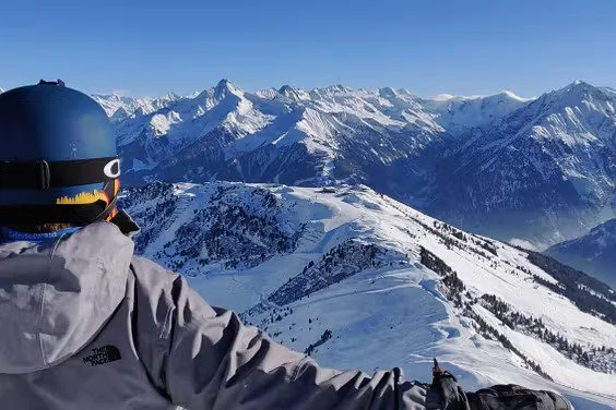 Person wearing a helmet and grey jacket standing on a snowy mountain ridge overlooking snow-capped mountains and peaks.