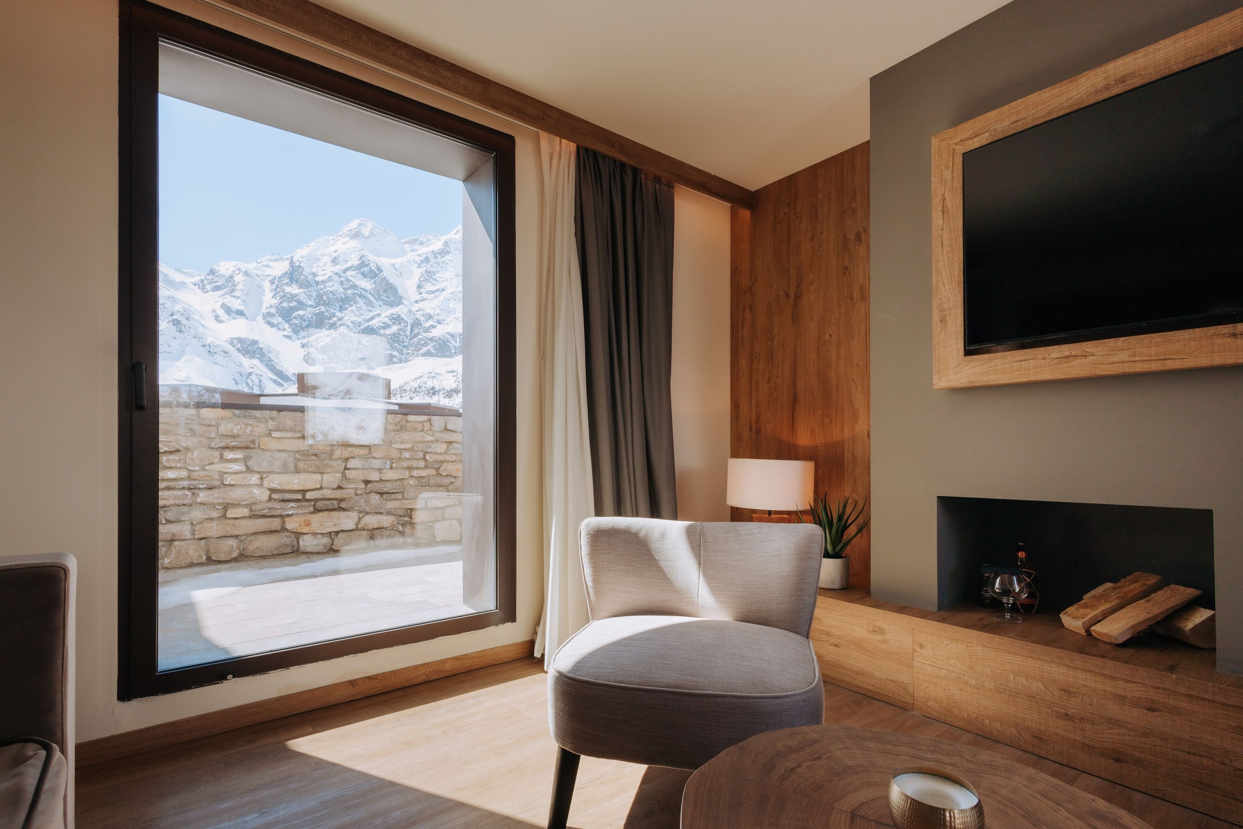 Living room with large window showing snow-covered mountains and stone balcony, beige and wood decor, chair, lamp, and television.