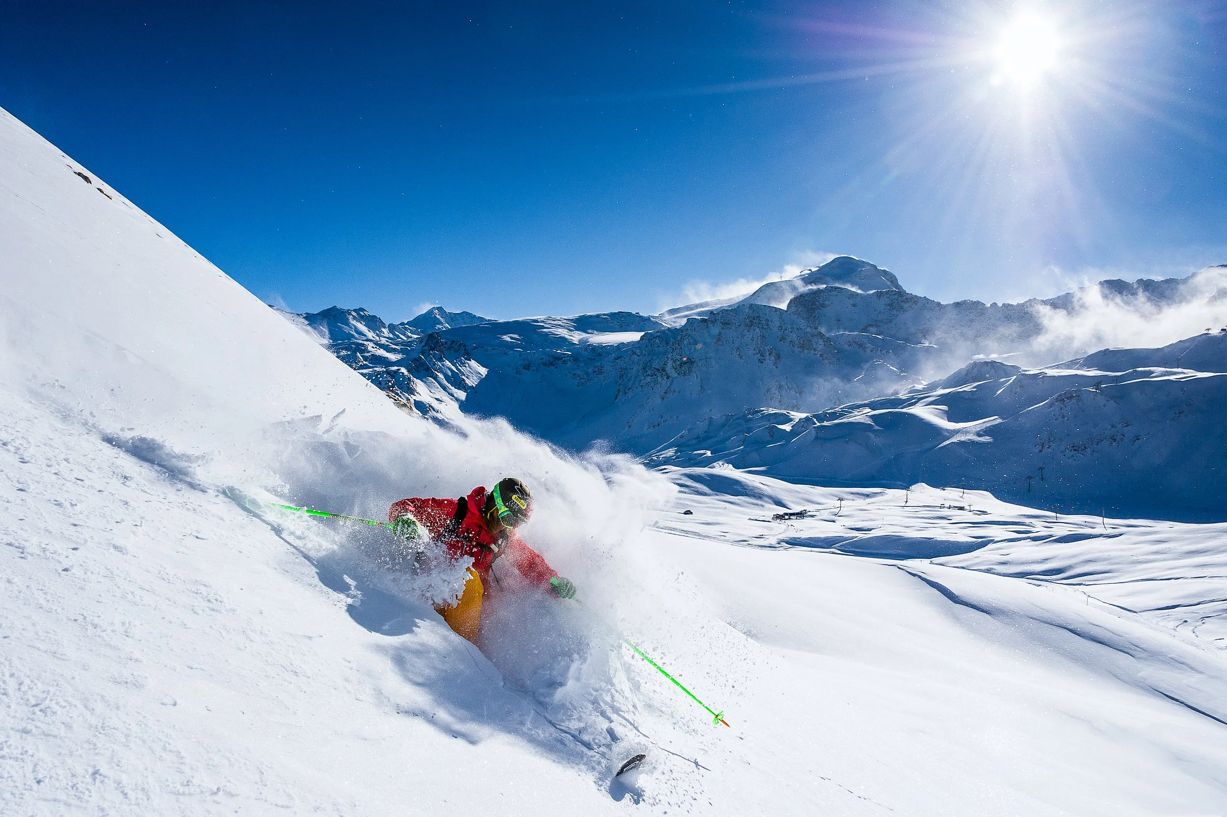 A skier wearing a red jacket and yellow pants skiing down a snowy mountain slope on a bright, sunny day with clear blue sky and snow-covered peaks in the background.