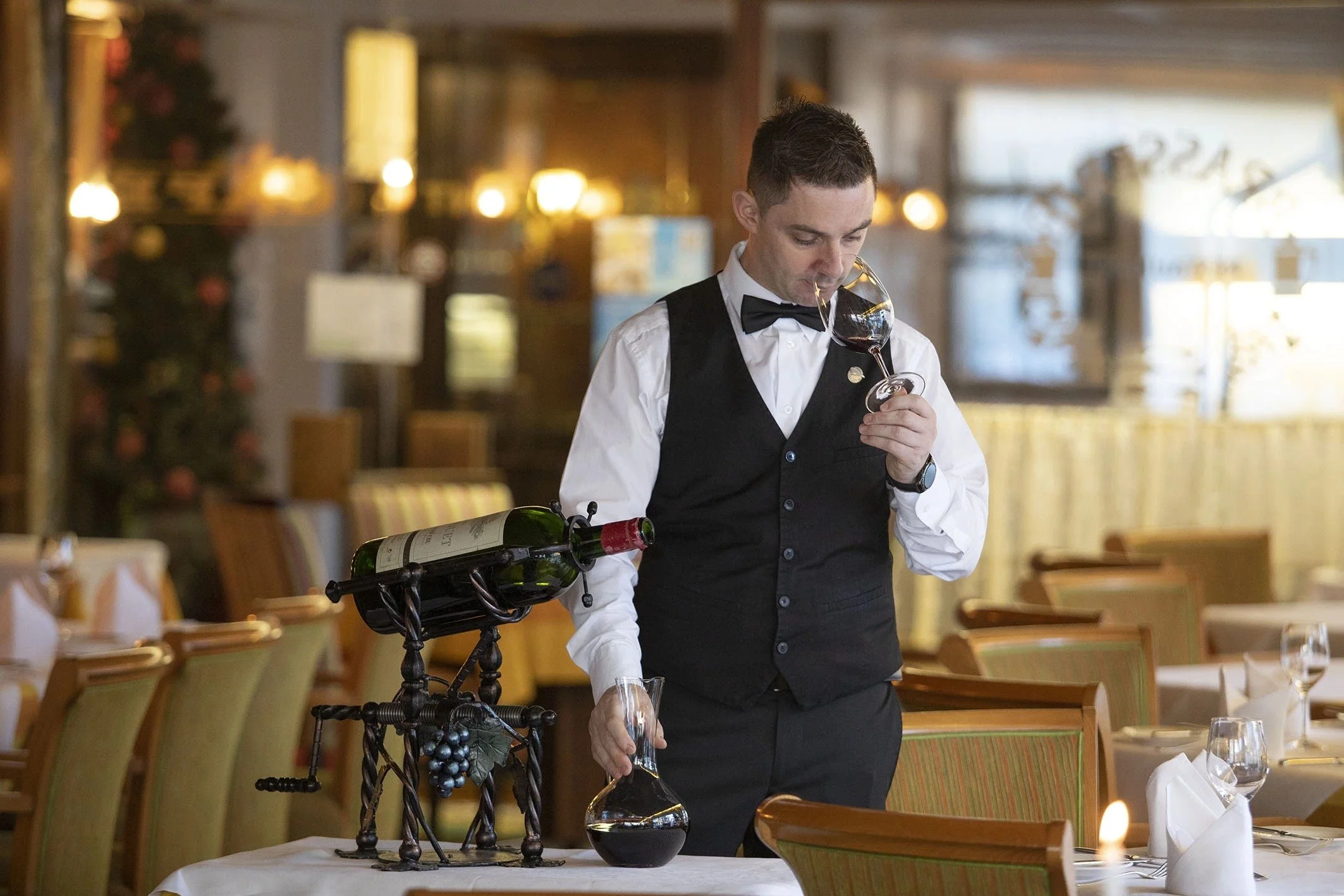 A waiter in a tuxedo pouring red wine from a bottle into a glass on a table in a restaurant.