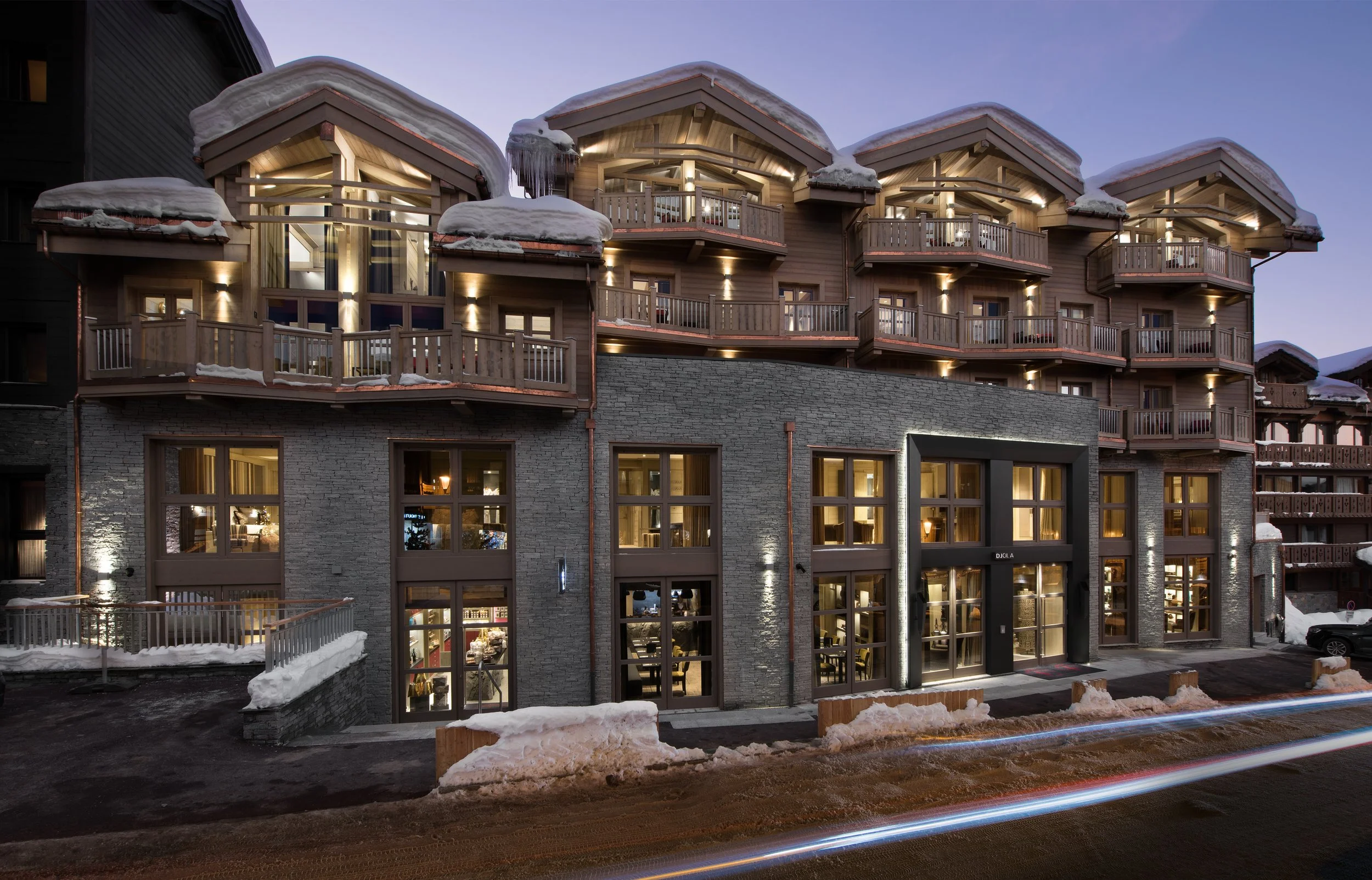 A multi-story mountain-style building with wooden balconies and large windows, illuminated at dusk, covered in snow and ice.
