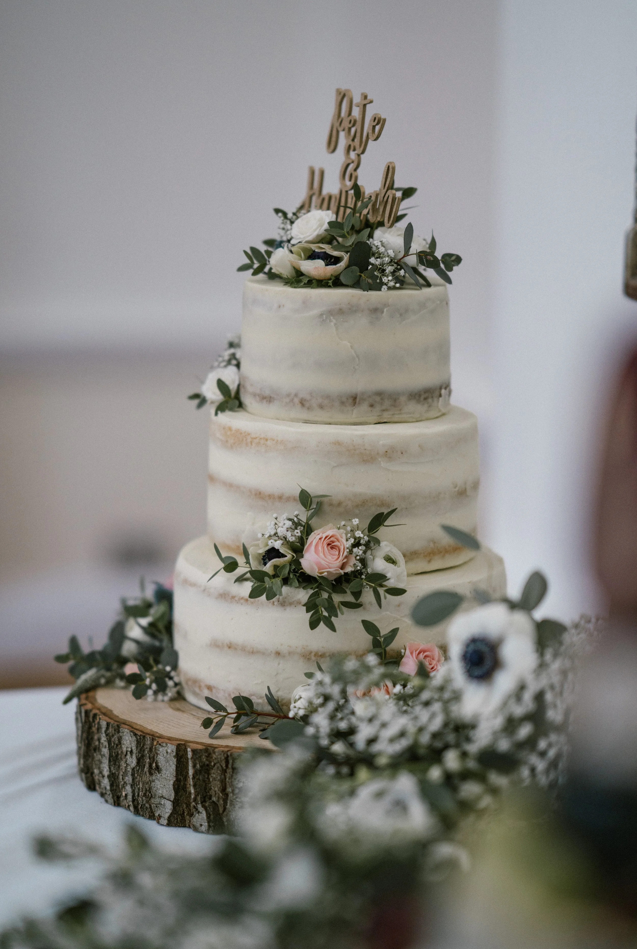Three-tiered wedding cake decorated with pink and white roses, white anemones, and greenery, with a gold 'Pete & Heather' cake topper, placed on a wooden slab with floral arrangements.