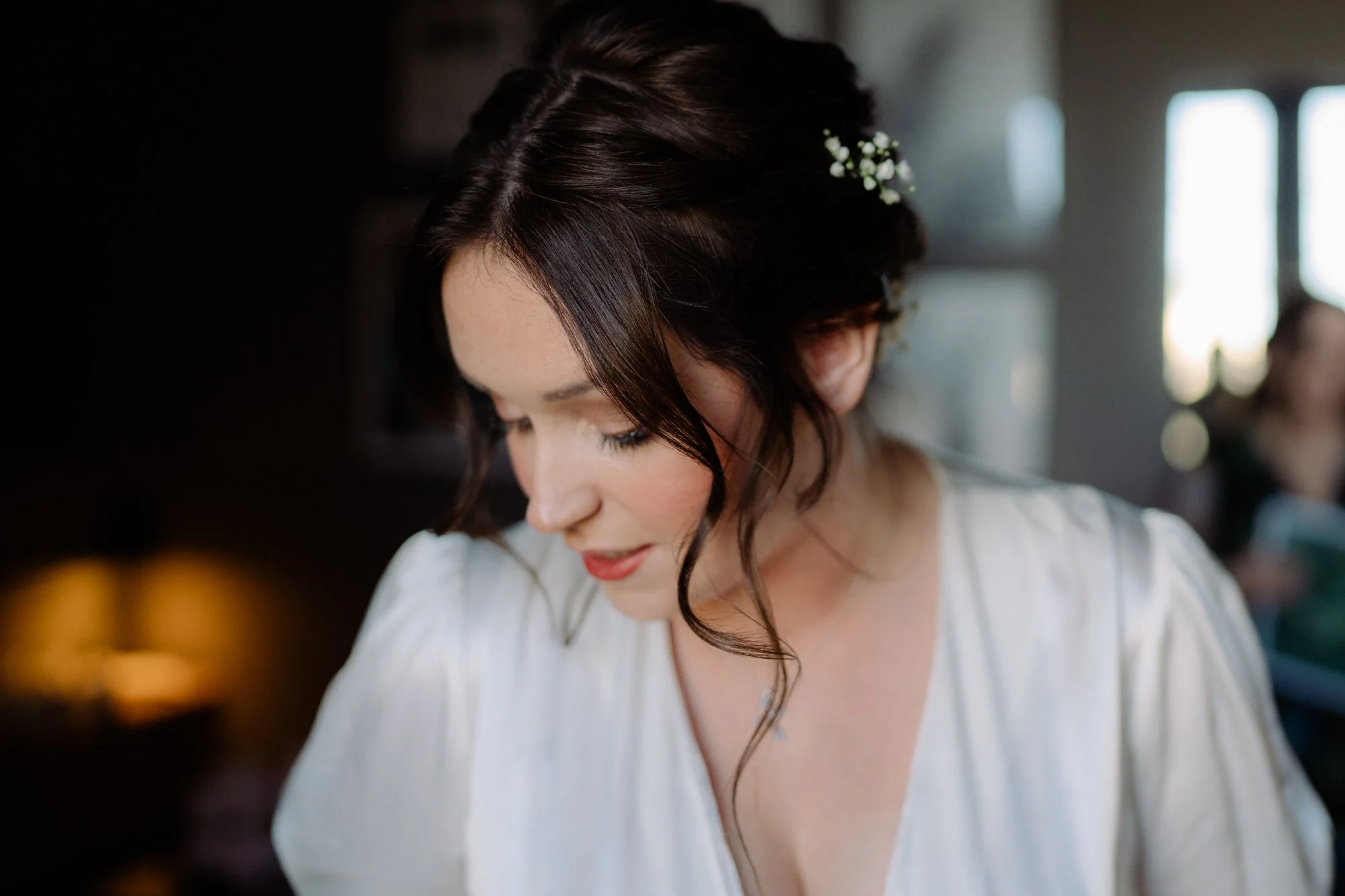 A woman with dark hair styled in loose waves, wearing a white dress with puffed sleeves, is looking down with her eyes closed. She has a small floral accessory in her hair and a delicate necklace, in a softly lit indoor setting.