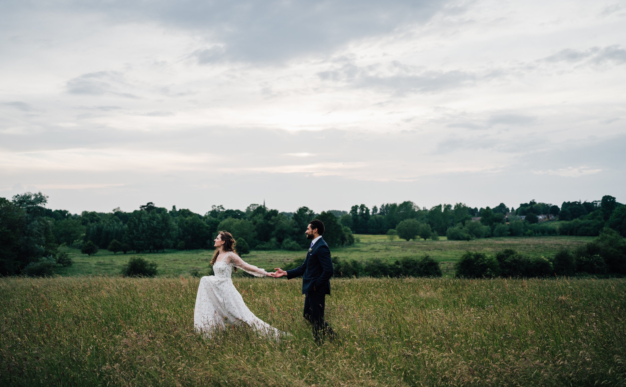 A newlywed couple holding hands in a grassy field with a cloudy sky and green trees in the background.
