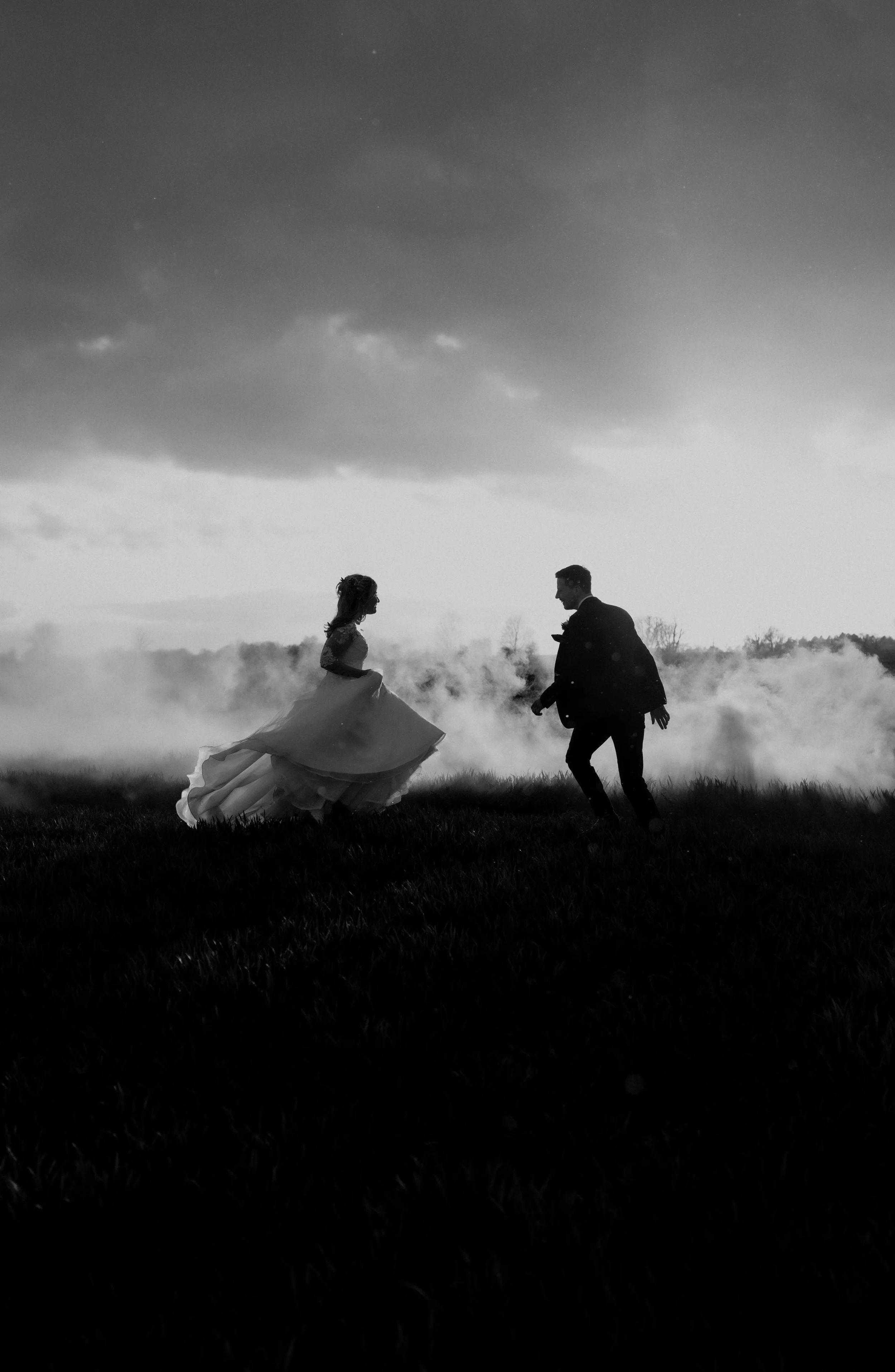 Black and white silhouette of a bride and groom dancing outdoors on a grassy field with cloudy sky and fog in the background.