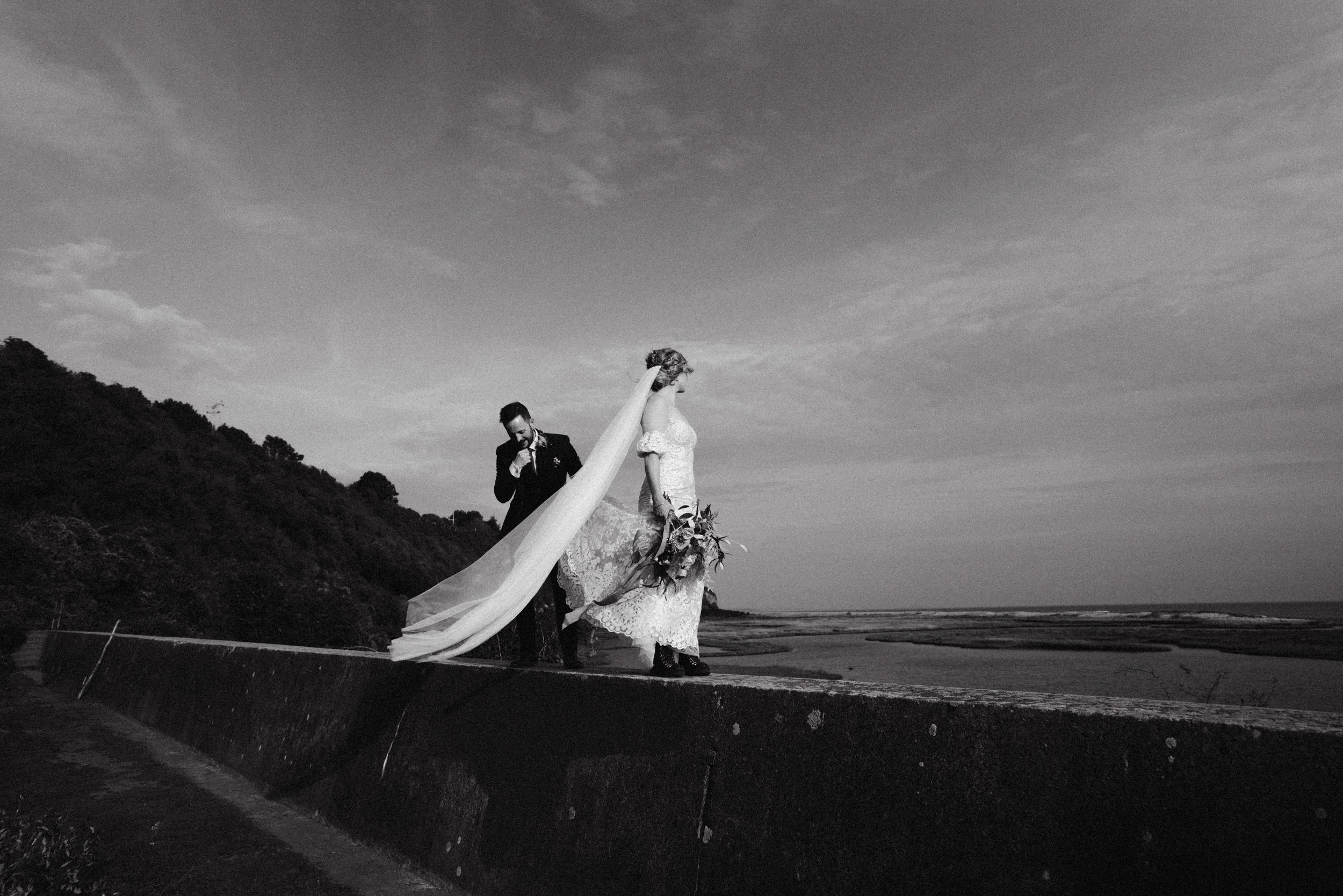A black and white photo of a bride and groom walking on a coastal wall, with the bride holding a bouquet and the groom adjusting his tie, against a backdrop of sky, sea, and hills.