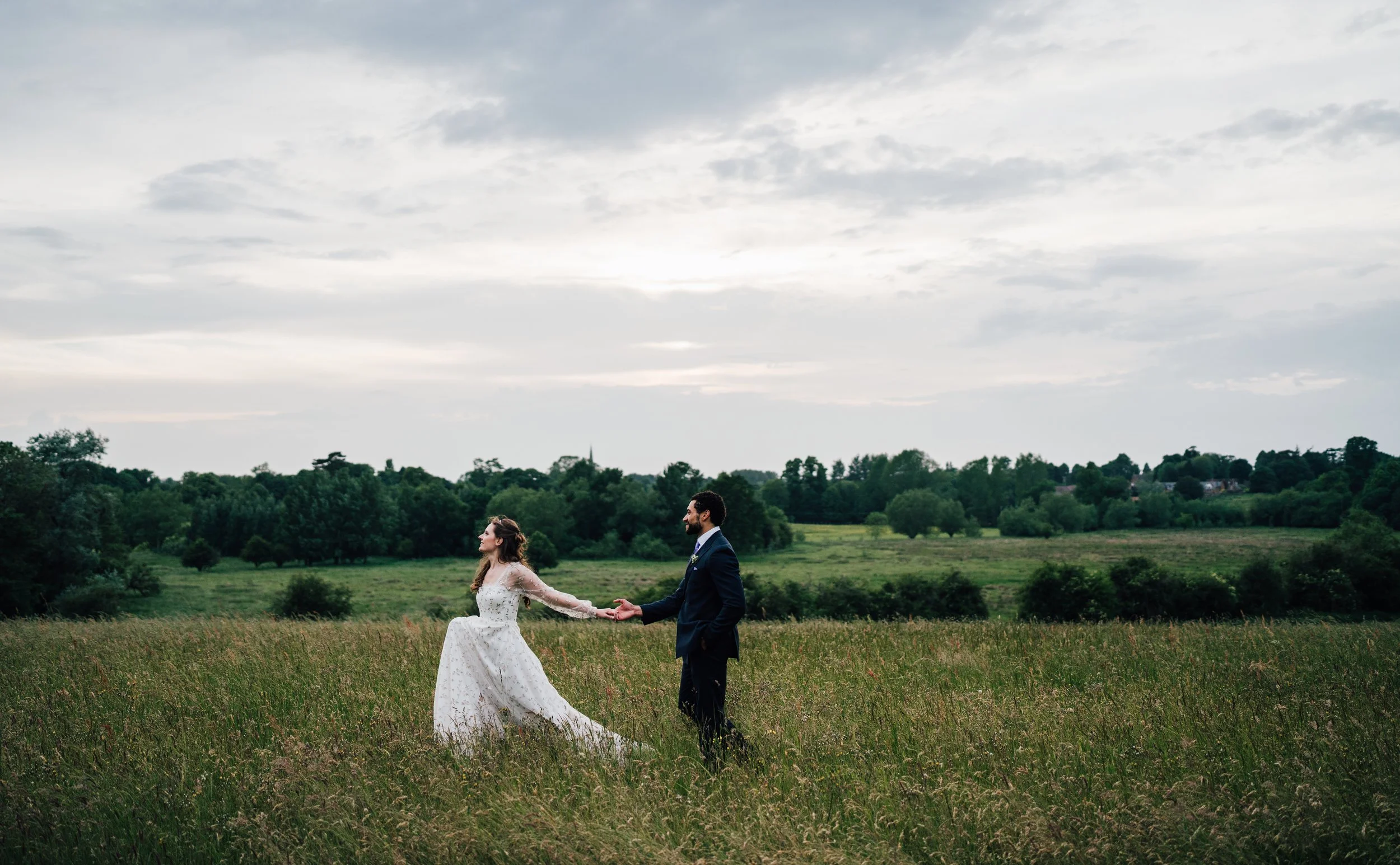 A bride and groom holding hands in a grassy field with trees and a cloudy sky in the background.