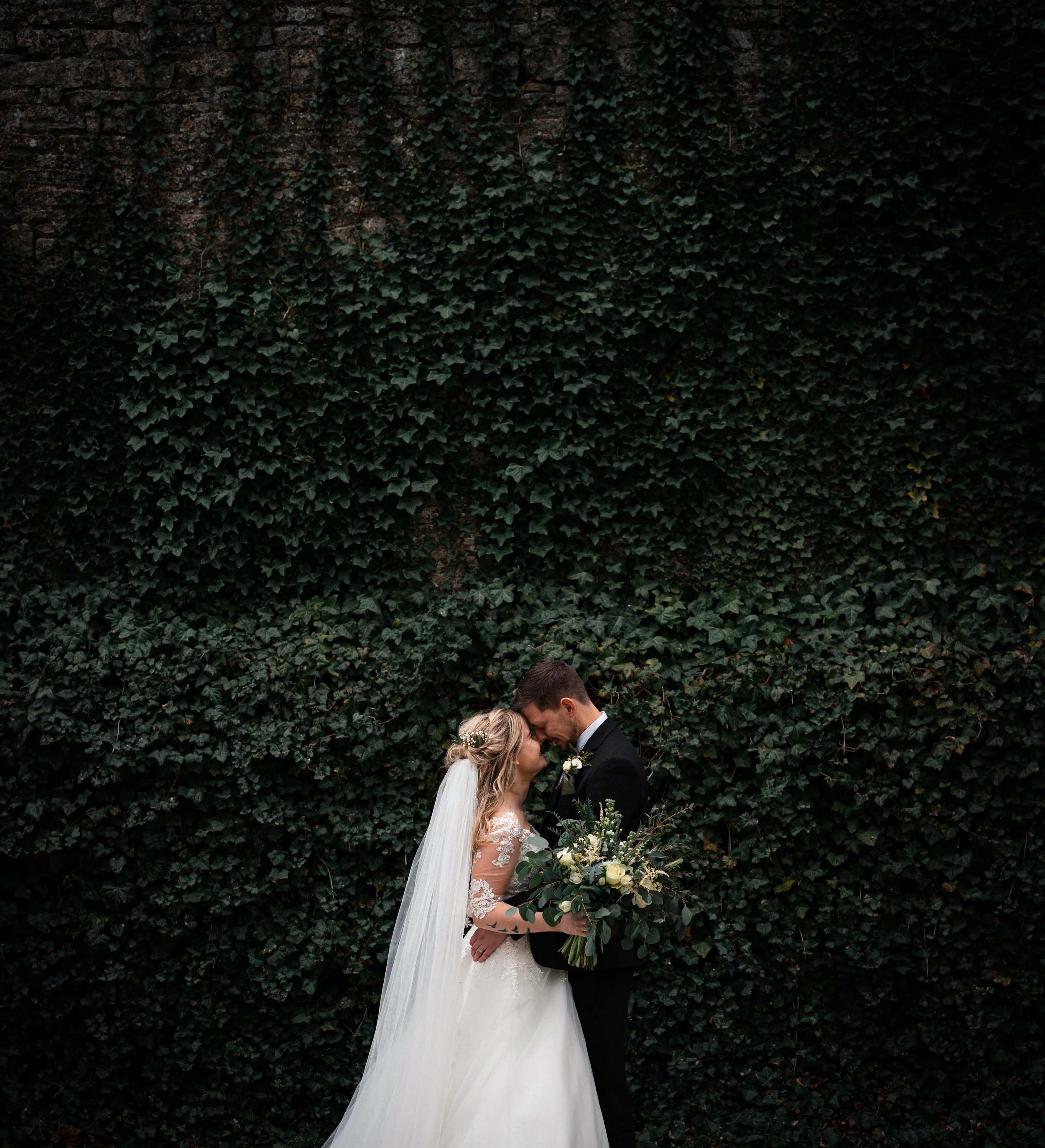 A bride in a white wedding dress and veil, and a groom in a black tuxedo, stand close together with foreheads touching in front of a dark green ivy-covered wall, holding a bouquet of white and green flowers.